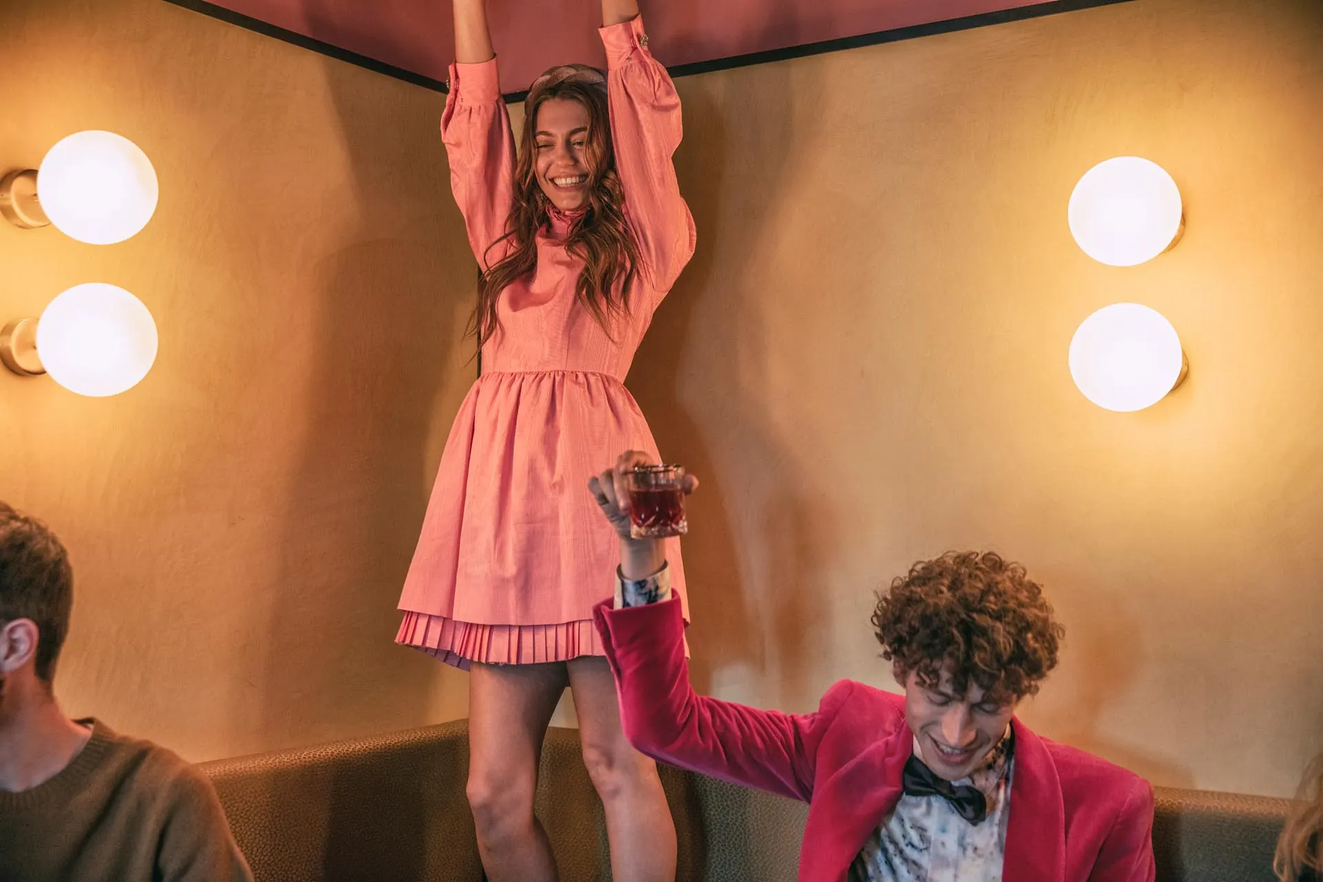 Smiling woman in pink dress stands with arms raised while man in bright suit holds a drink cheerfully.