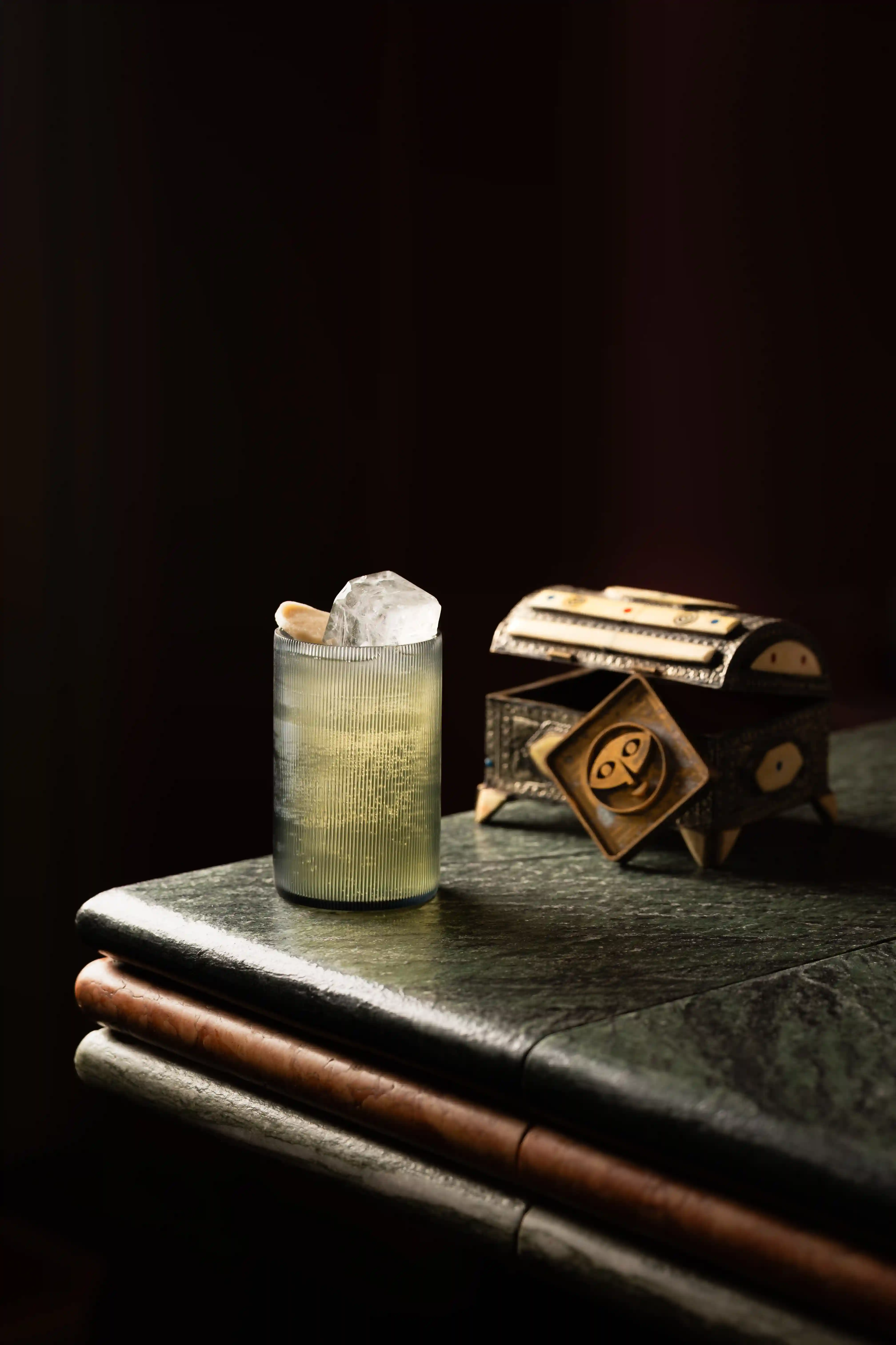 Glass with large ice cube next to an ornate small chest on green marble table.