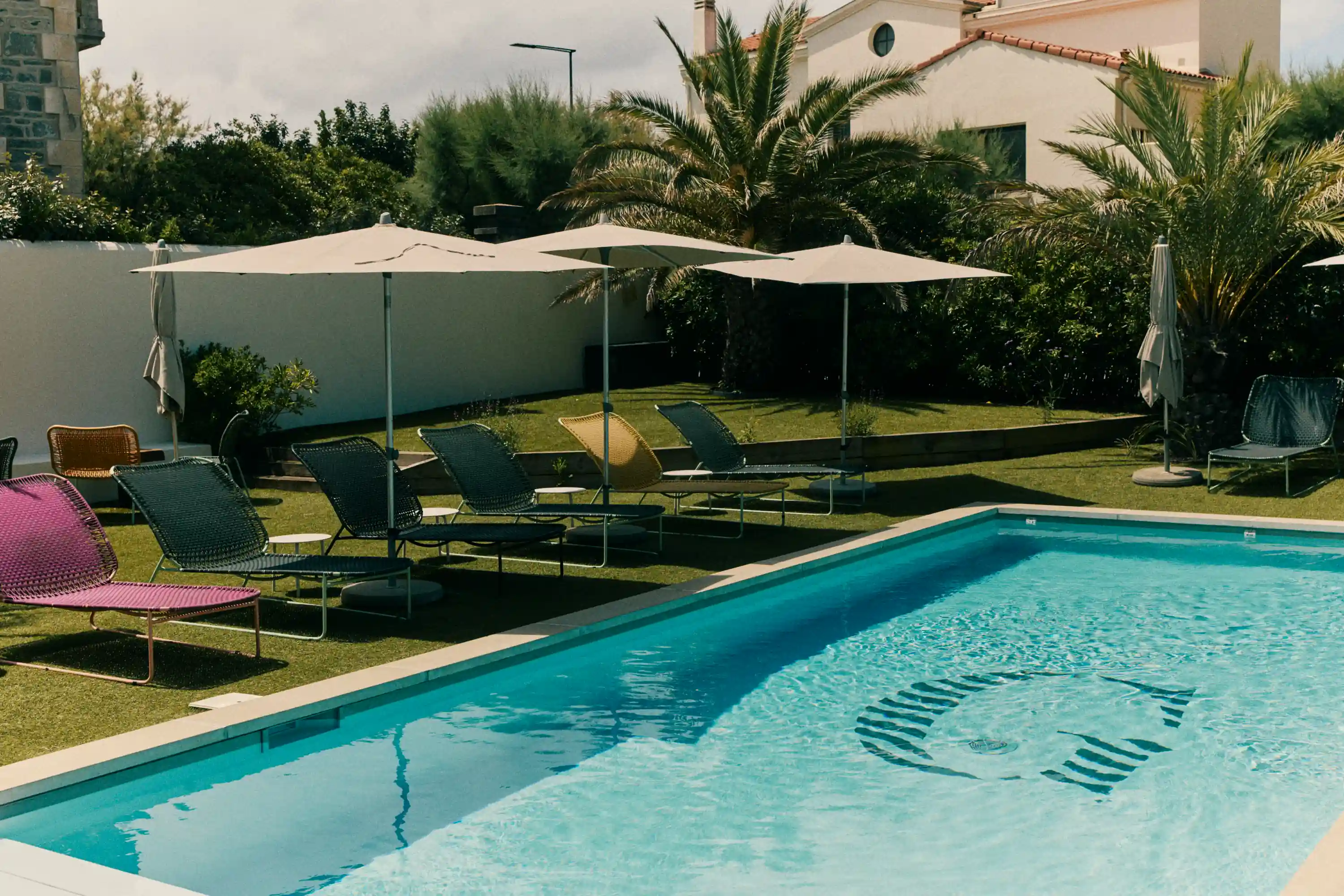 Outdoor pool with turquoise water, lounge chairs, white umbrellas, palm trees, and a house in the background.