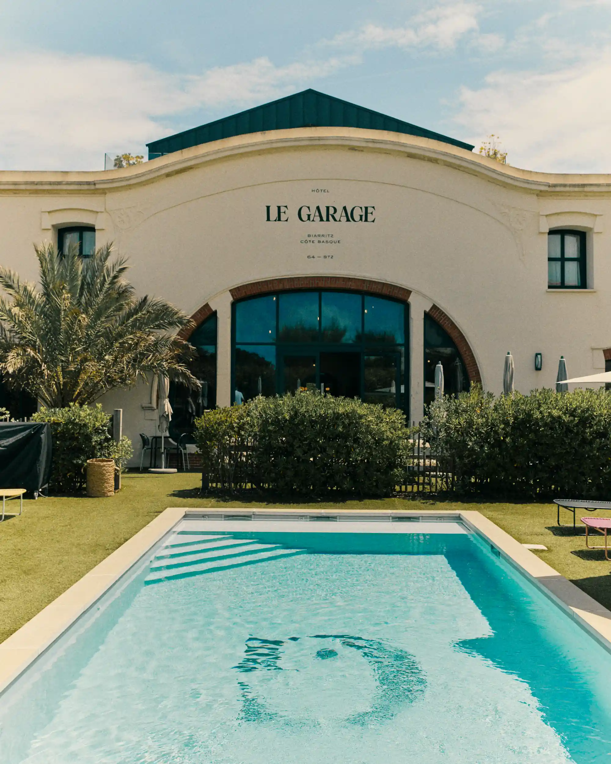 Outdoor pool with clear blue water in front of Le Garage hotel building with greenery and palm trees.