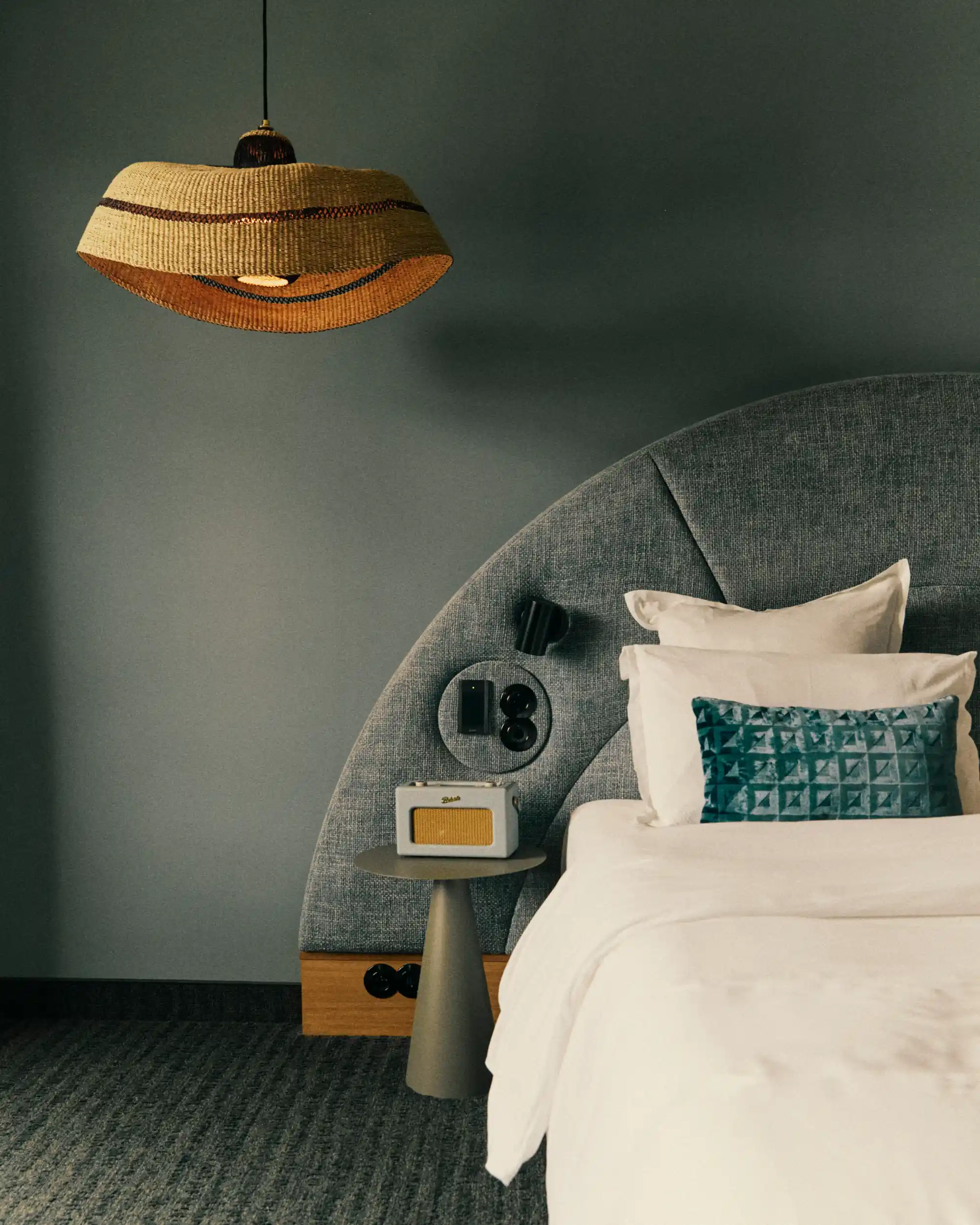 Modern bedroom corner with rounded gray headboard, bedside table, vintage radio, and hanging woven lamp.