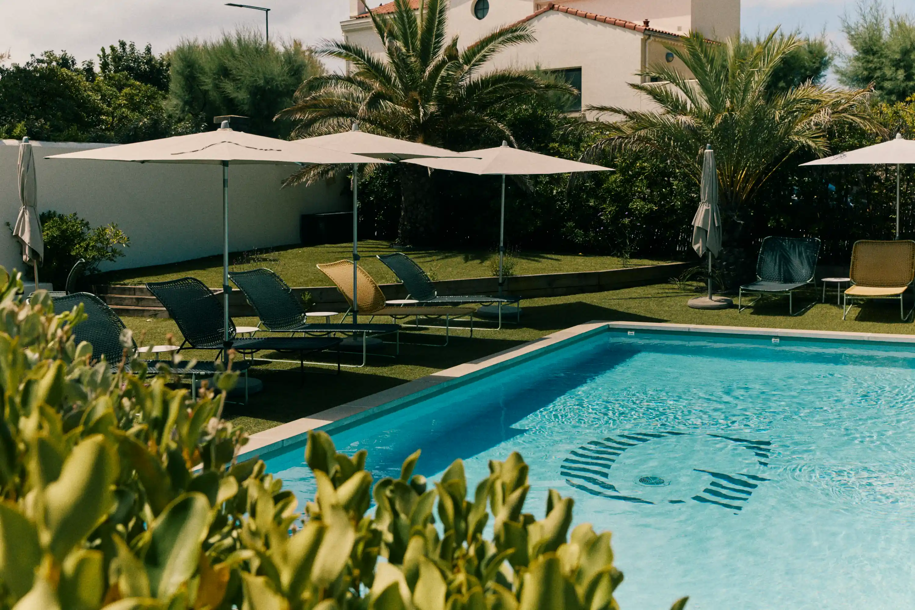 Outdoor pool with clear blue water, lounge chairs, umbrellas, palm trees, and a house in the background.