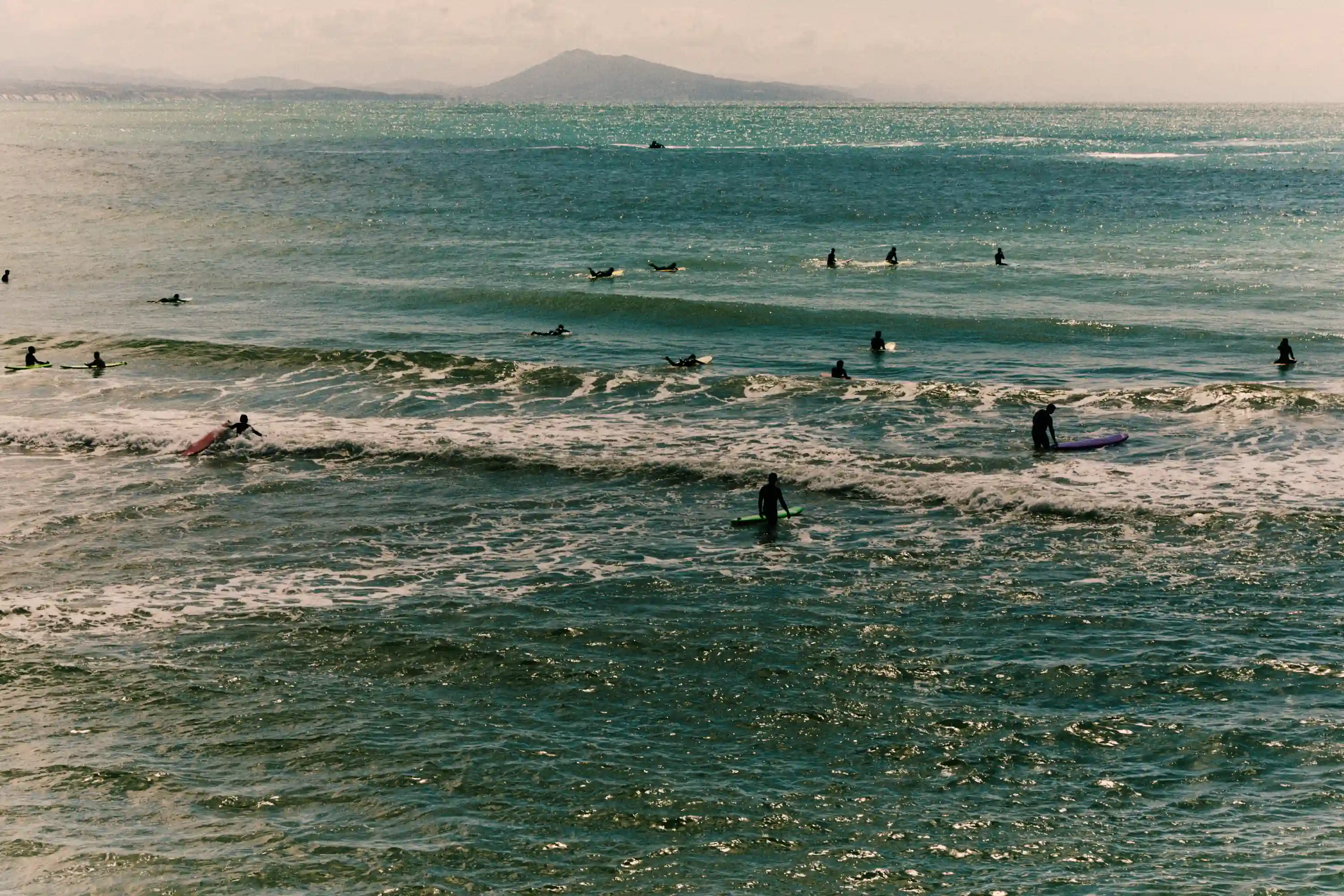 Surfers in wetsuits on surfboards in the ocean with a misty mountain in the background.