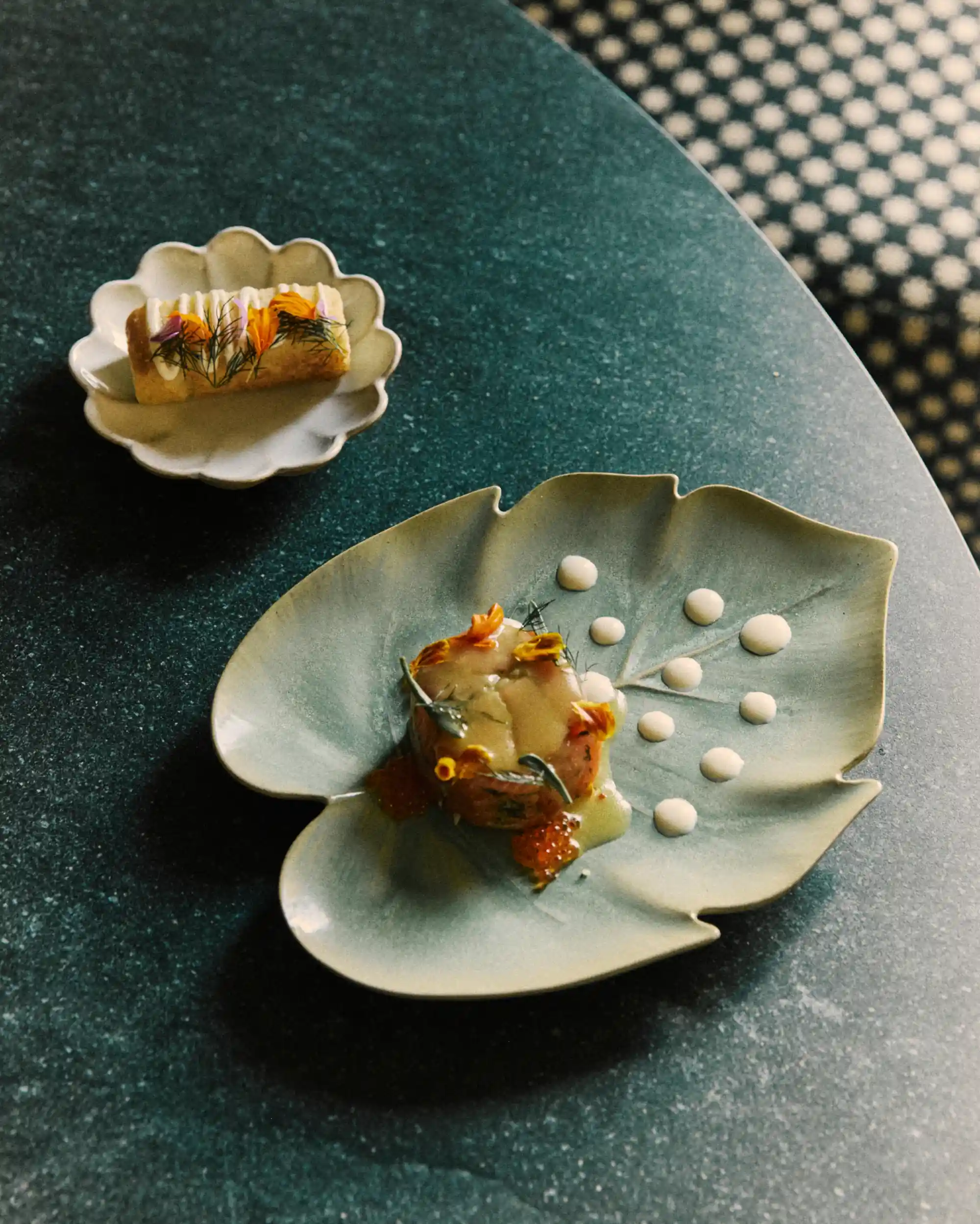 Two small, artistic dishes on a dark green table: one on a leaf plate, one on a scalloped plate.
