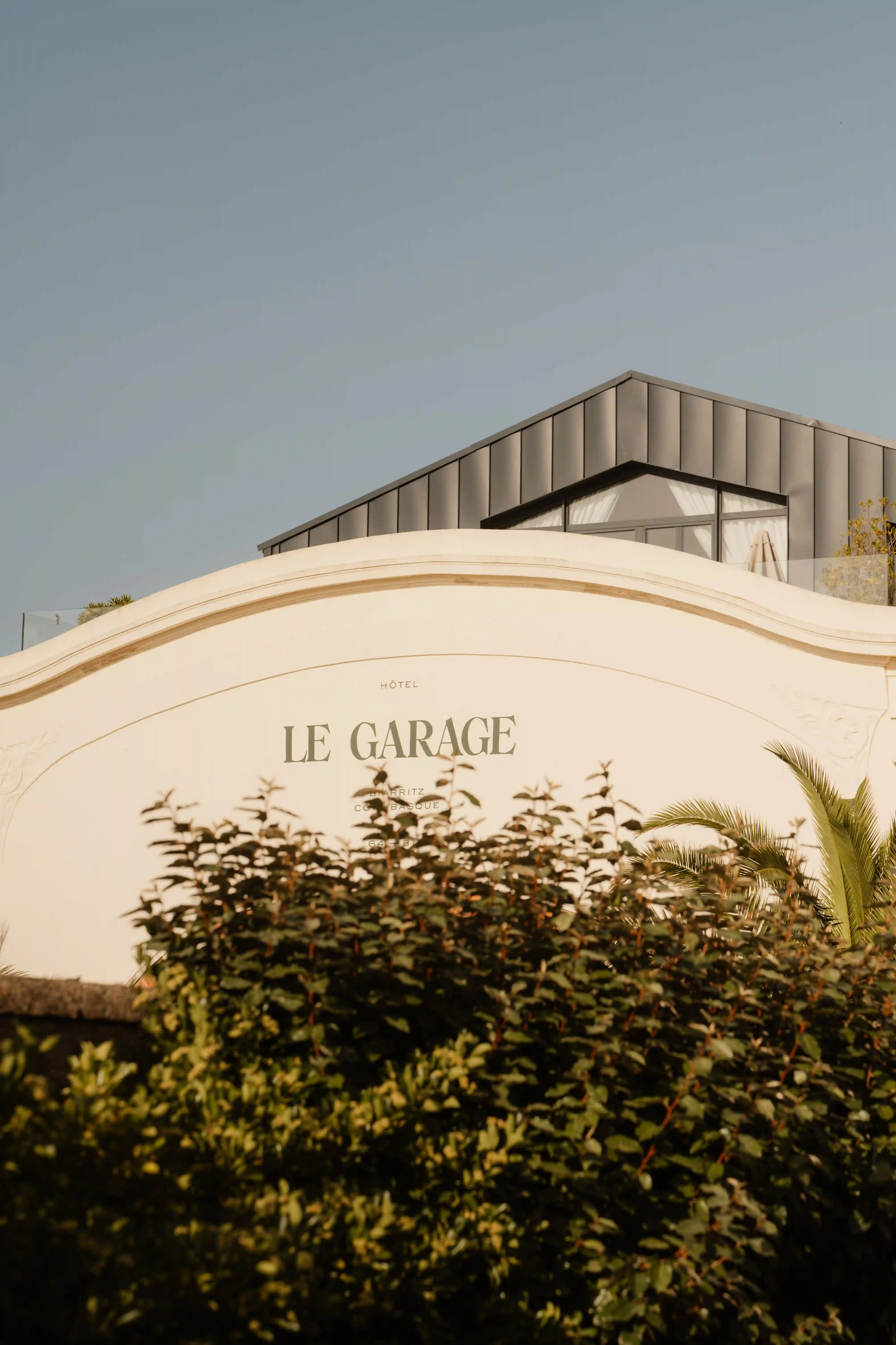 Curved white wall with "Le Garage" sign partially hidden by green bushes under a modern rooftop.
