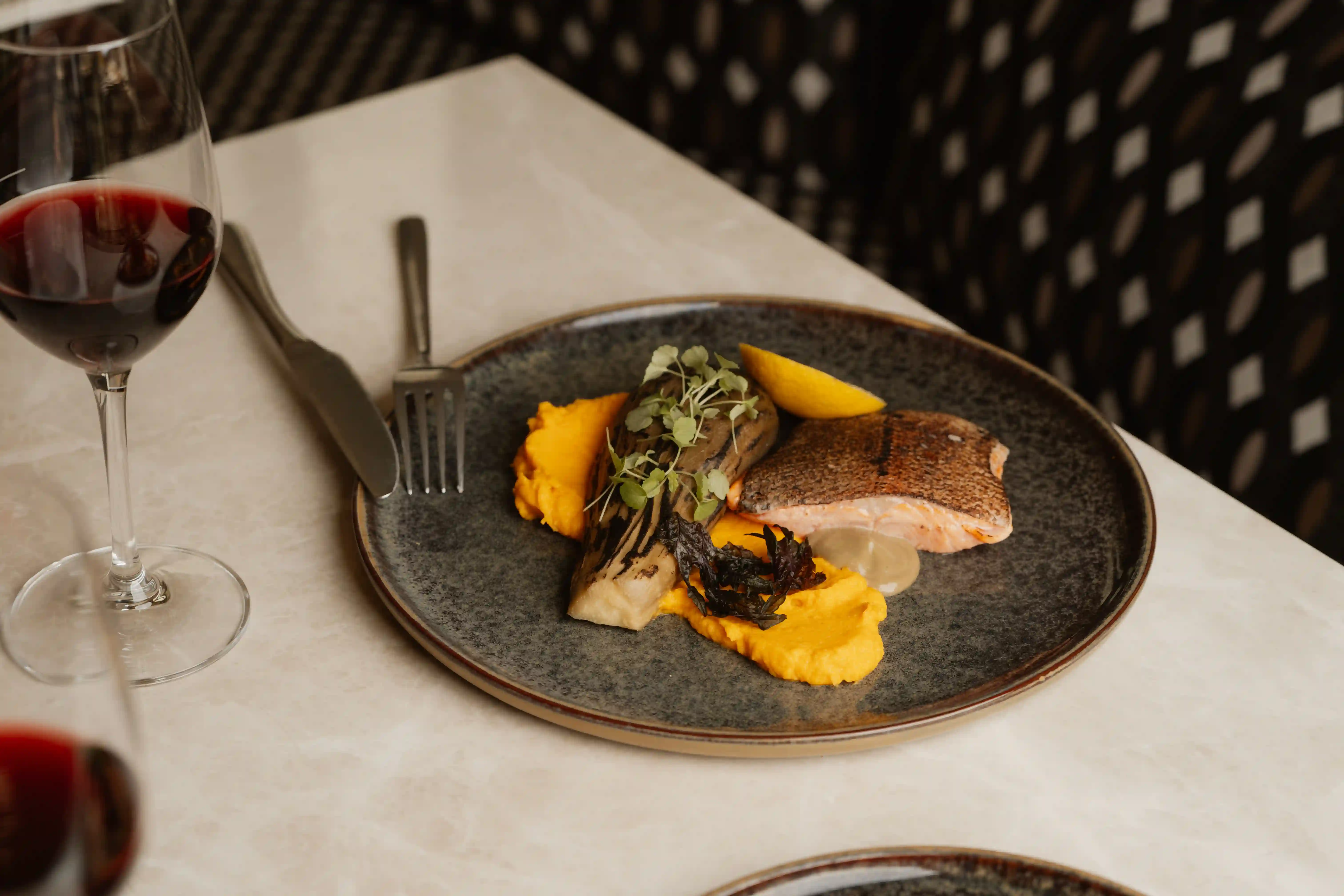 Plate with grilled salmon, orange puree, charred vegetable, greens, and lemon wedge on a table with wine.