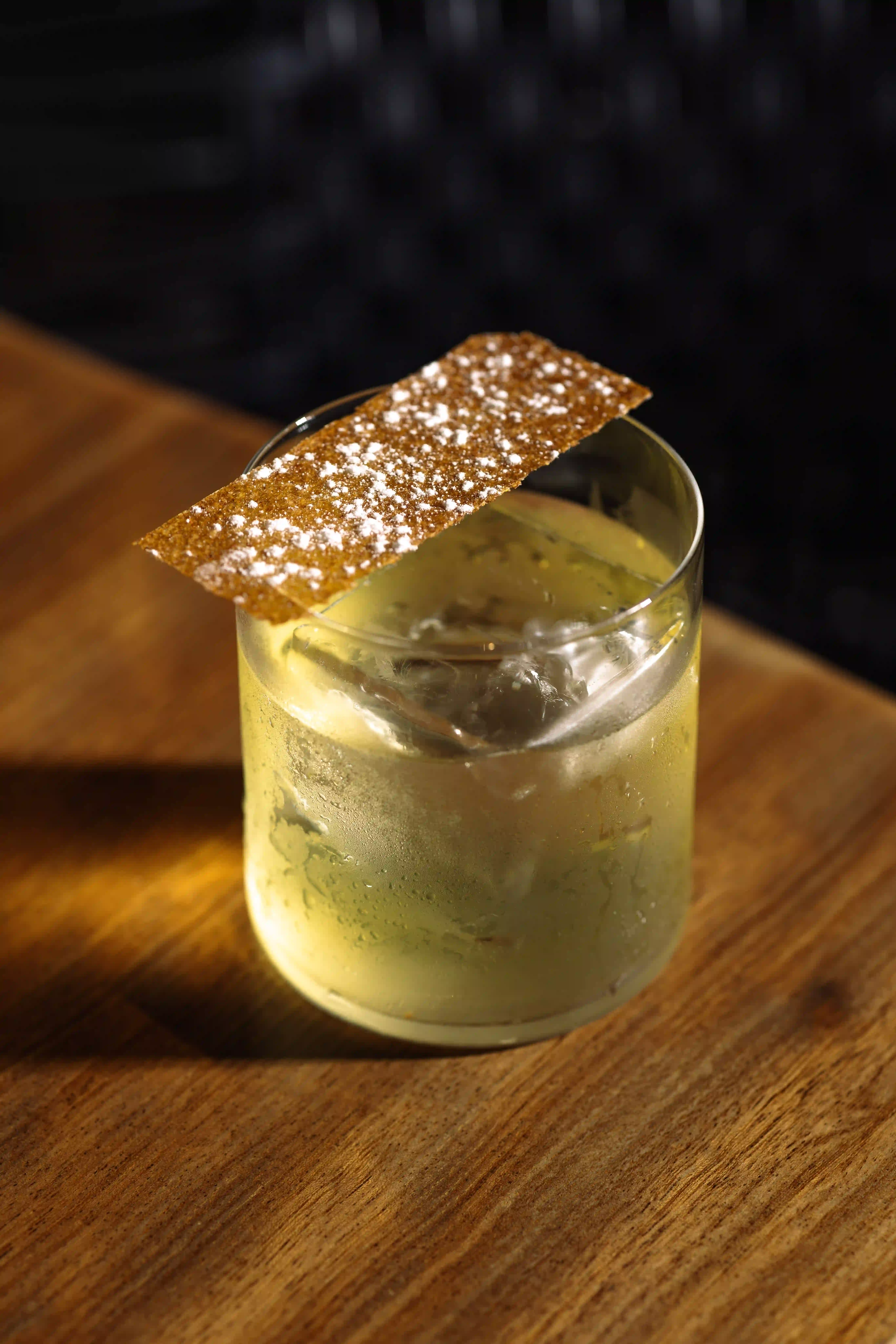 Clear glass with a light yellow cocktail, ice cubes, and a rectangular garnish sprinkled with white powder.