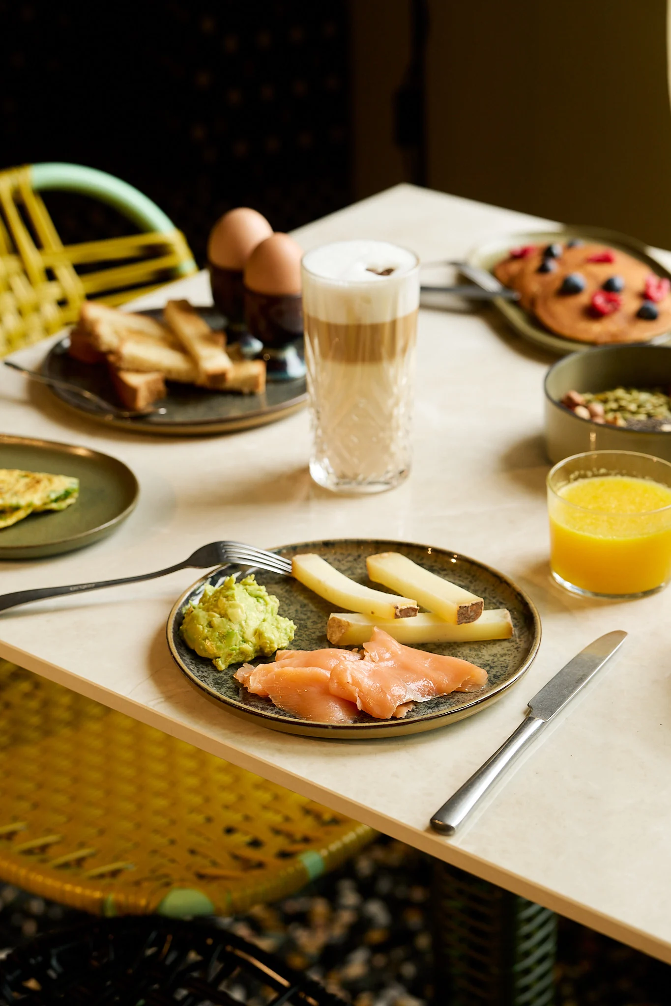 Table with breakfast including smoked salmon, avocado, cheese sticks, boiled eggs, toast, pancakes, orange juice, and latte.