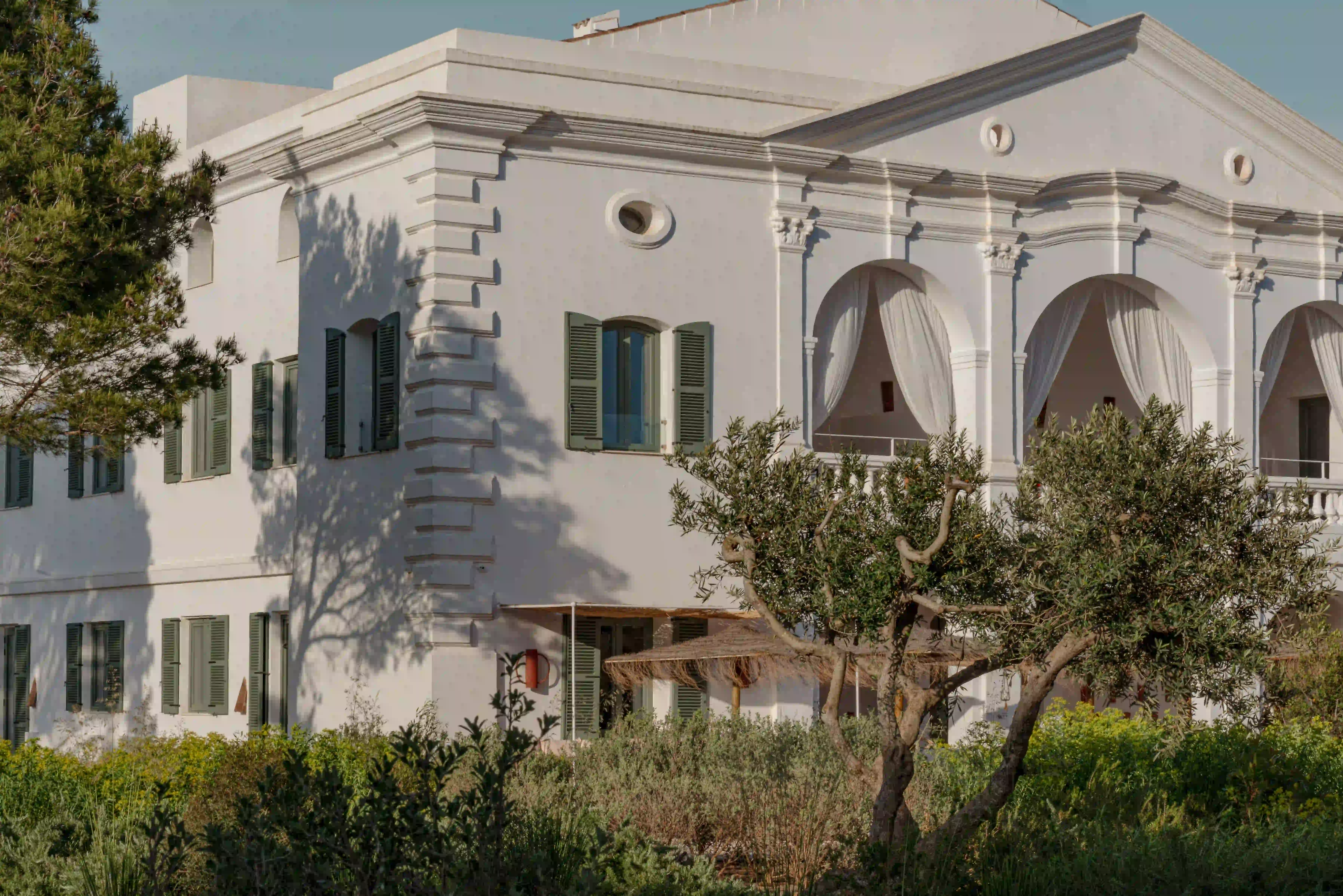 White Mediterranean villa with green shutters, arch balconies with curtains, surrounded by trees and shrubs.