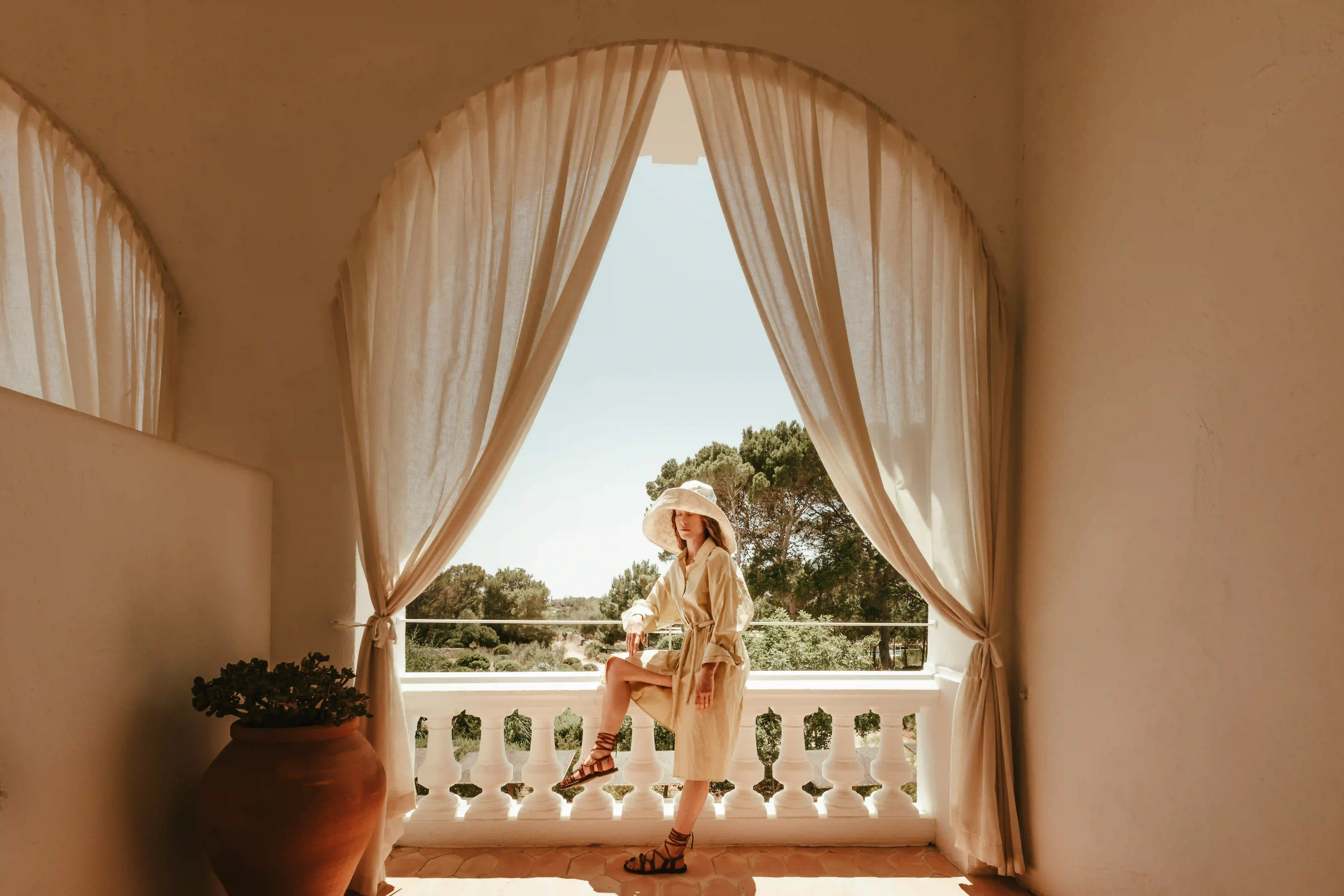 Woman in beige dress and wide hat posed on balcony with white curtains and greenery outside.
