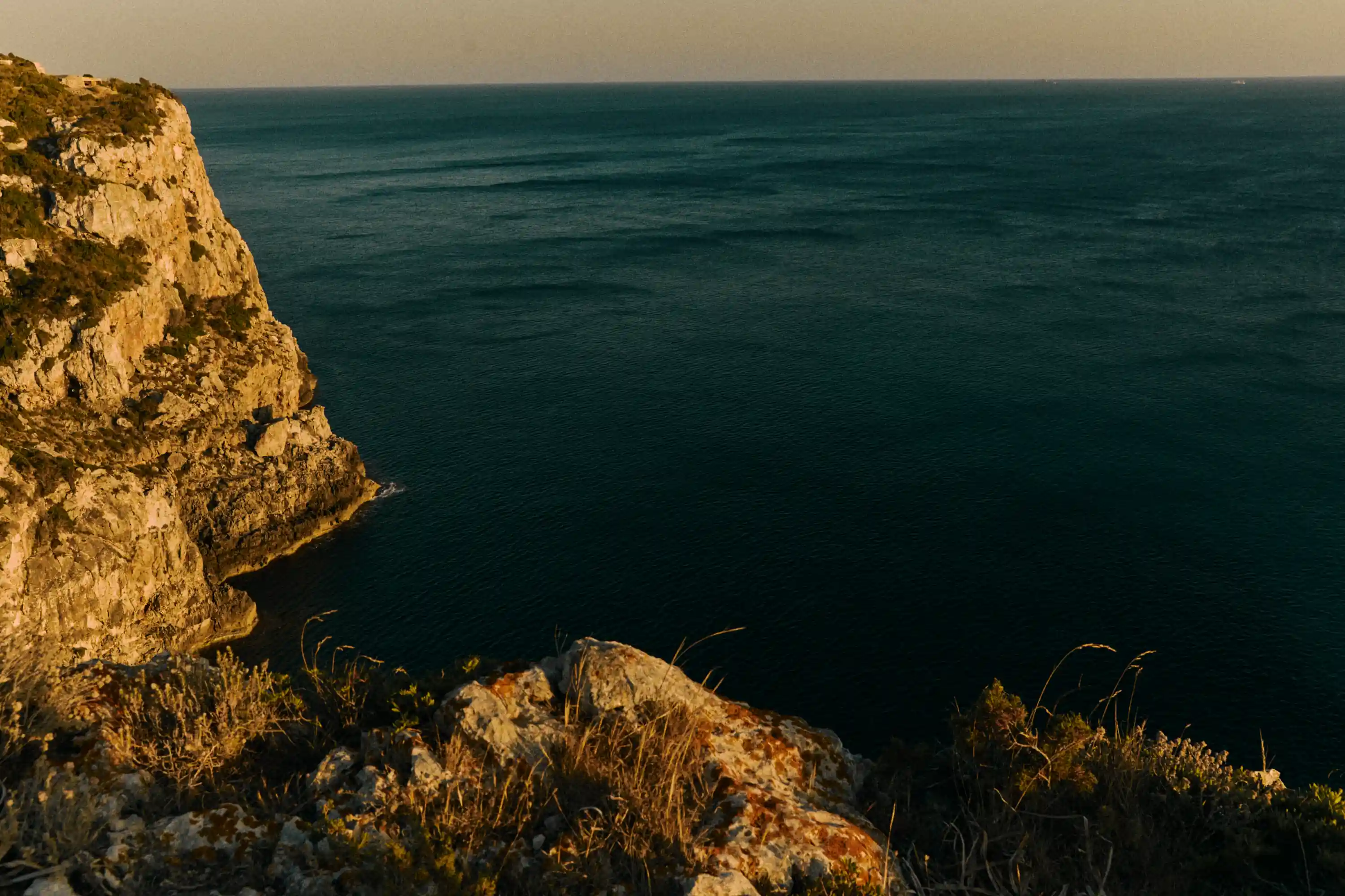 Rocky cliff edge with sparse grass overlooking calm dark blue ocean under clear sky.