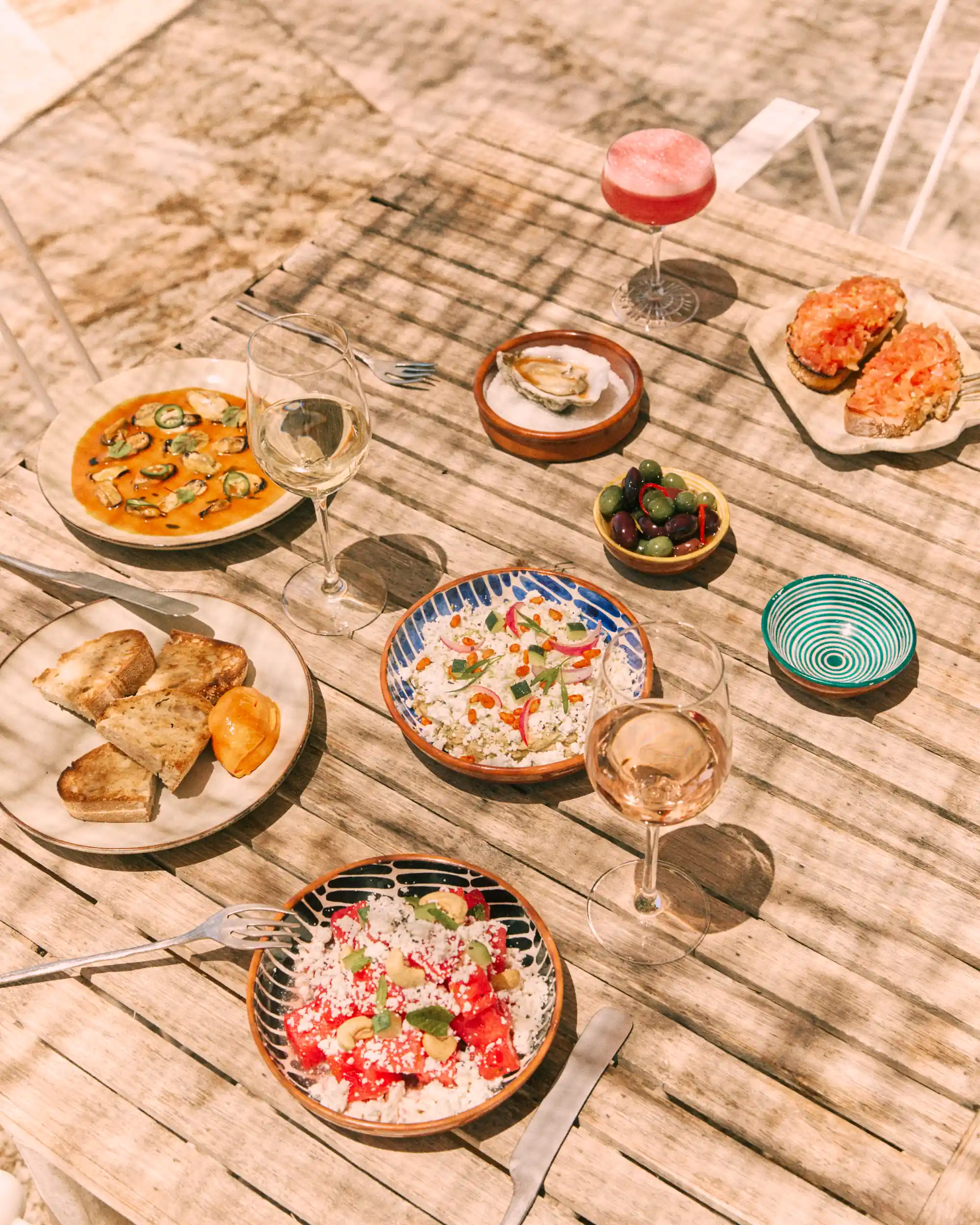 Wooden table with various dishes including bread, olives, oysters, salads, and wine glasses with drinks.