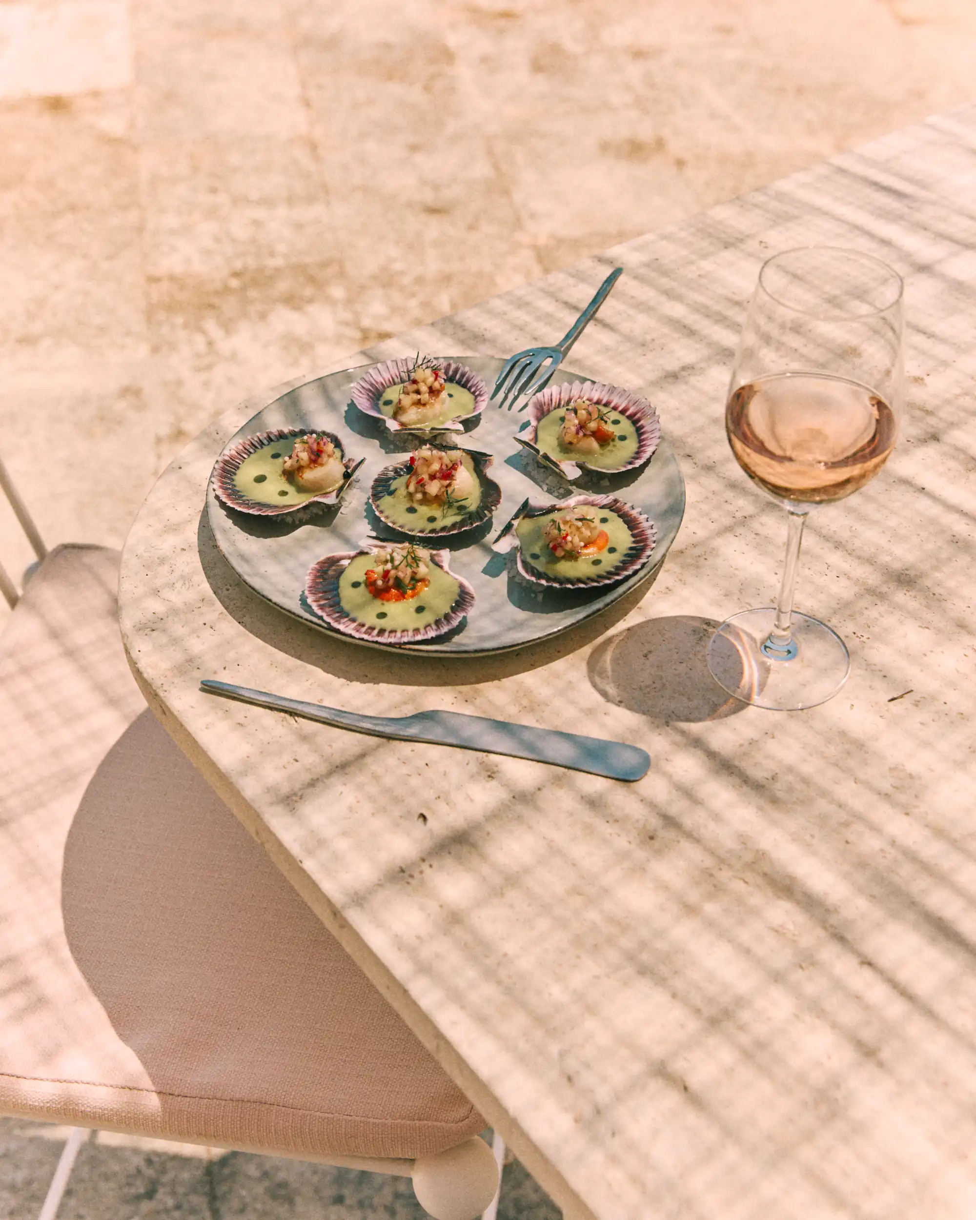 Plate with six scallops in shells topped with sauce and garnish beside a glass of rosé wine