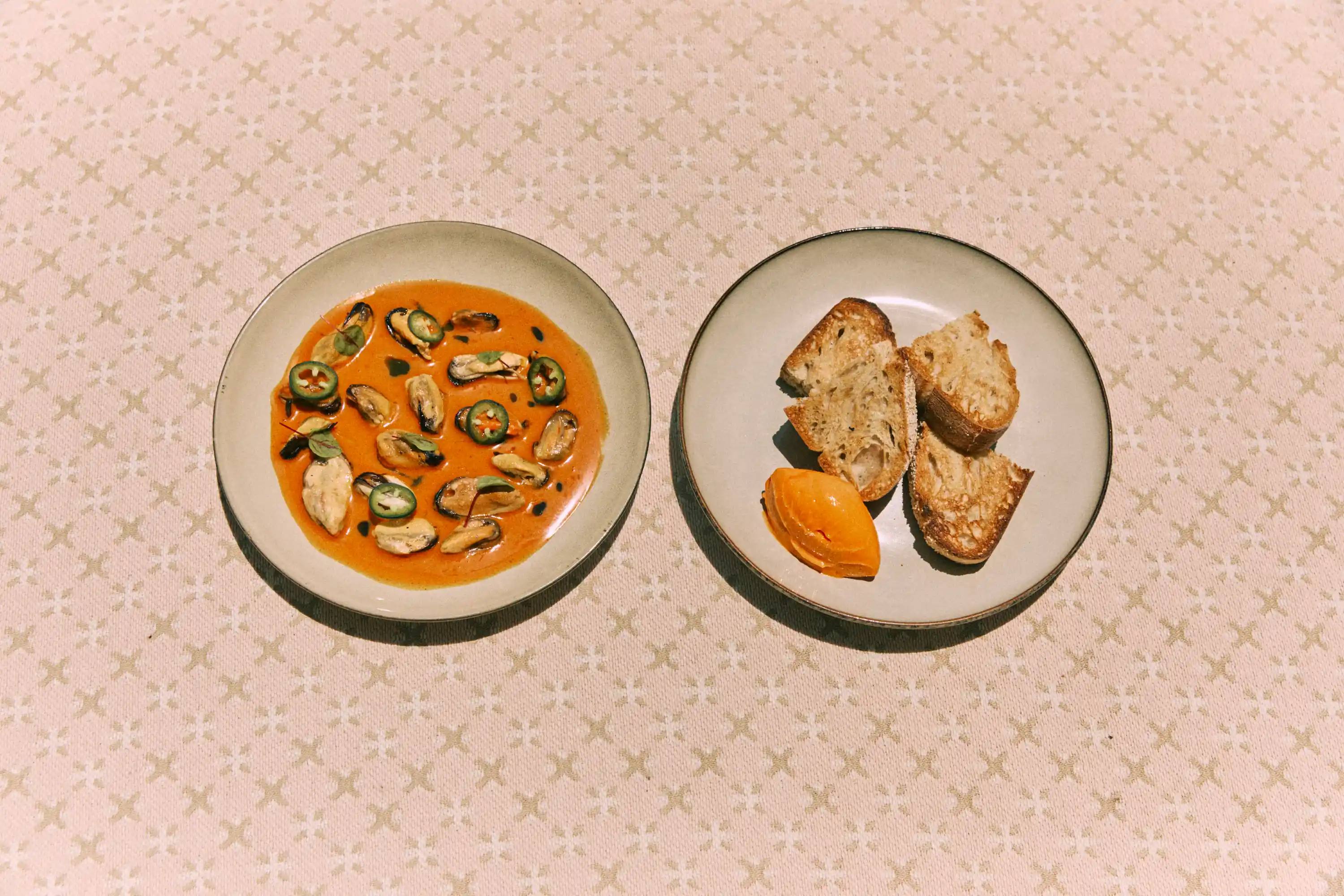 Two plates on a patterned cloth: one with seafood in sauce, the other with toasted bread and orange spread.