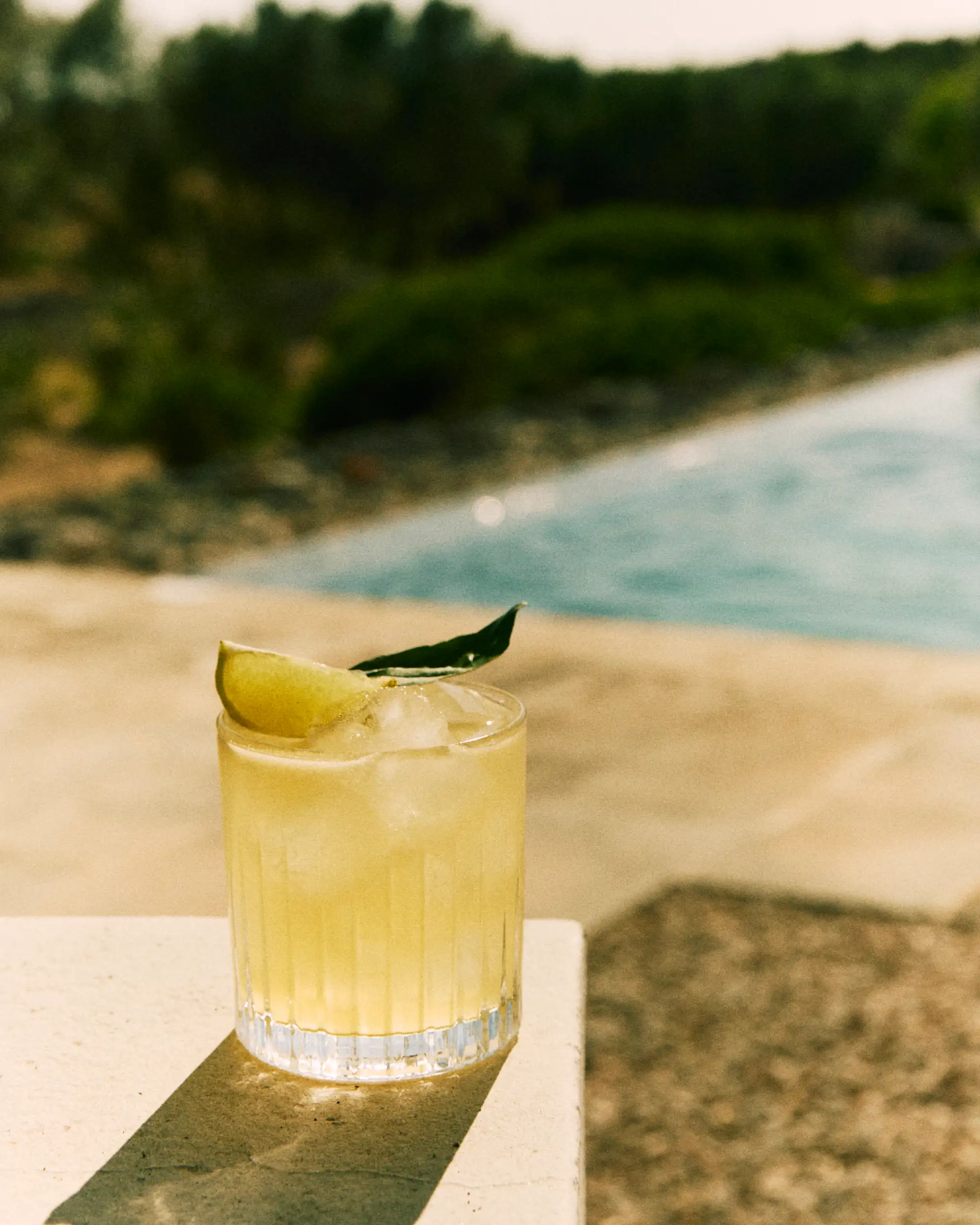 Yellow iced drink garnished with lime wedge and leaf by a pool on a sunny day.