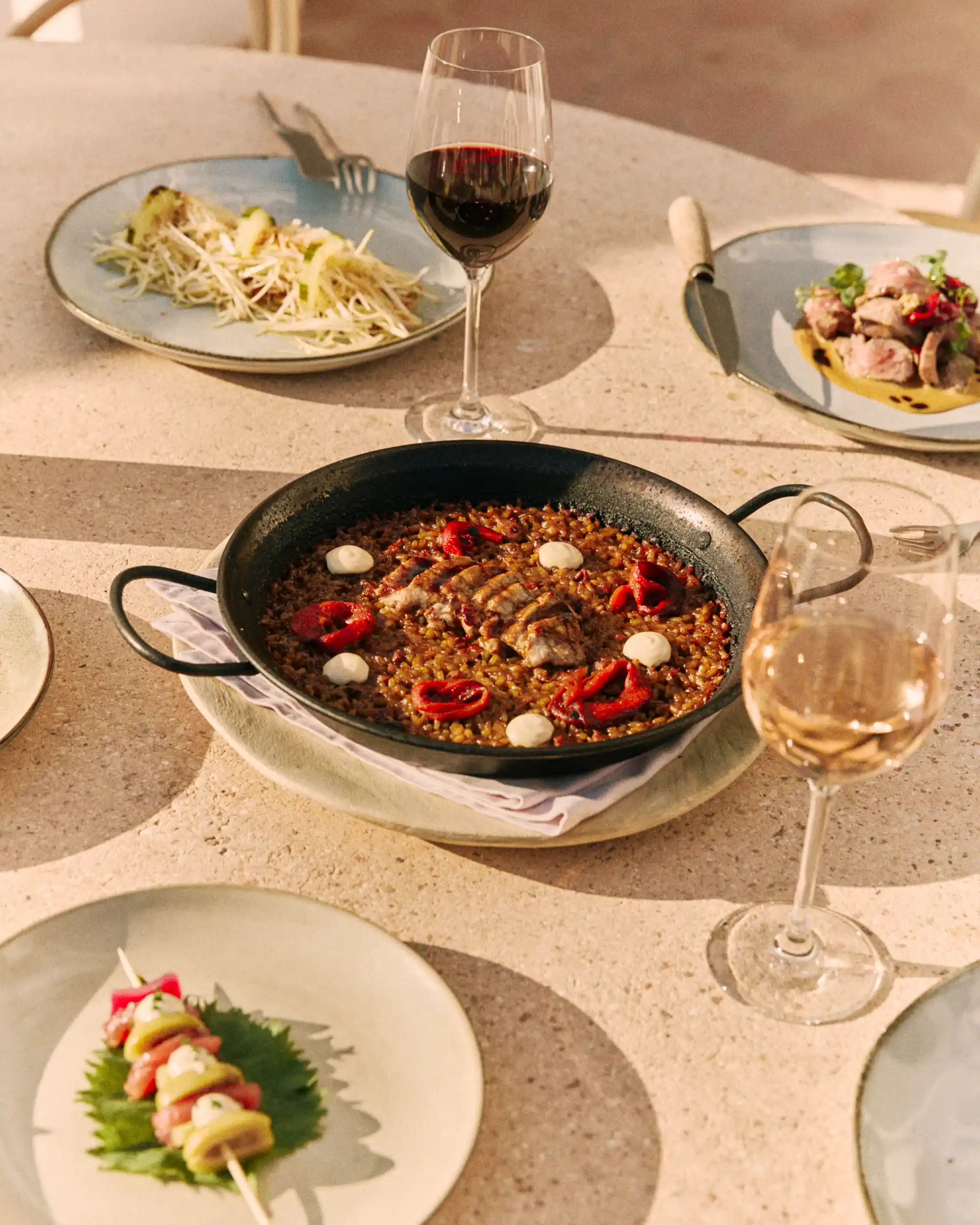 Table with a pan of rice dish garnished with red peppers, several plates of food, and two glasses of wine.