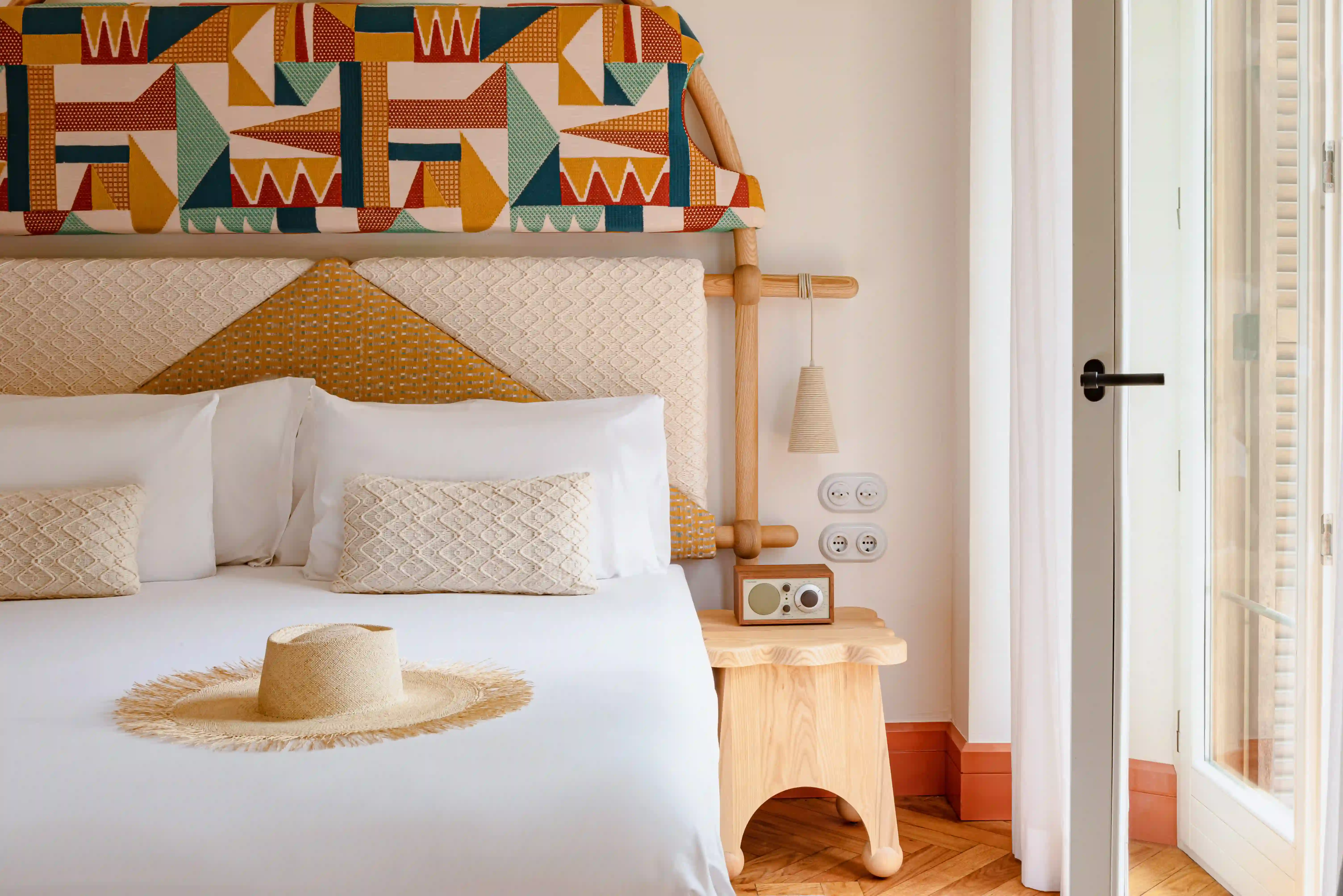 Bed with white linens, textured cushions, a straw hat, and a wooden nightstand with a retro radio.