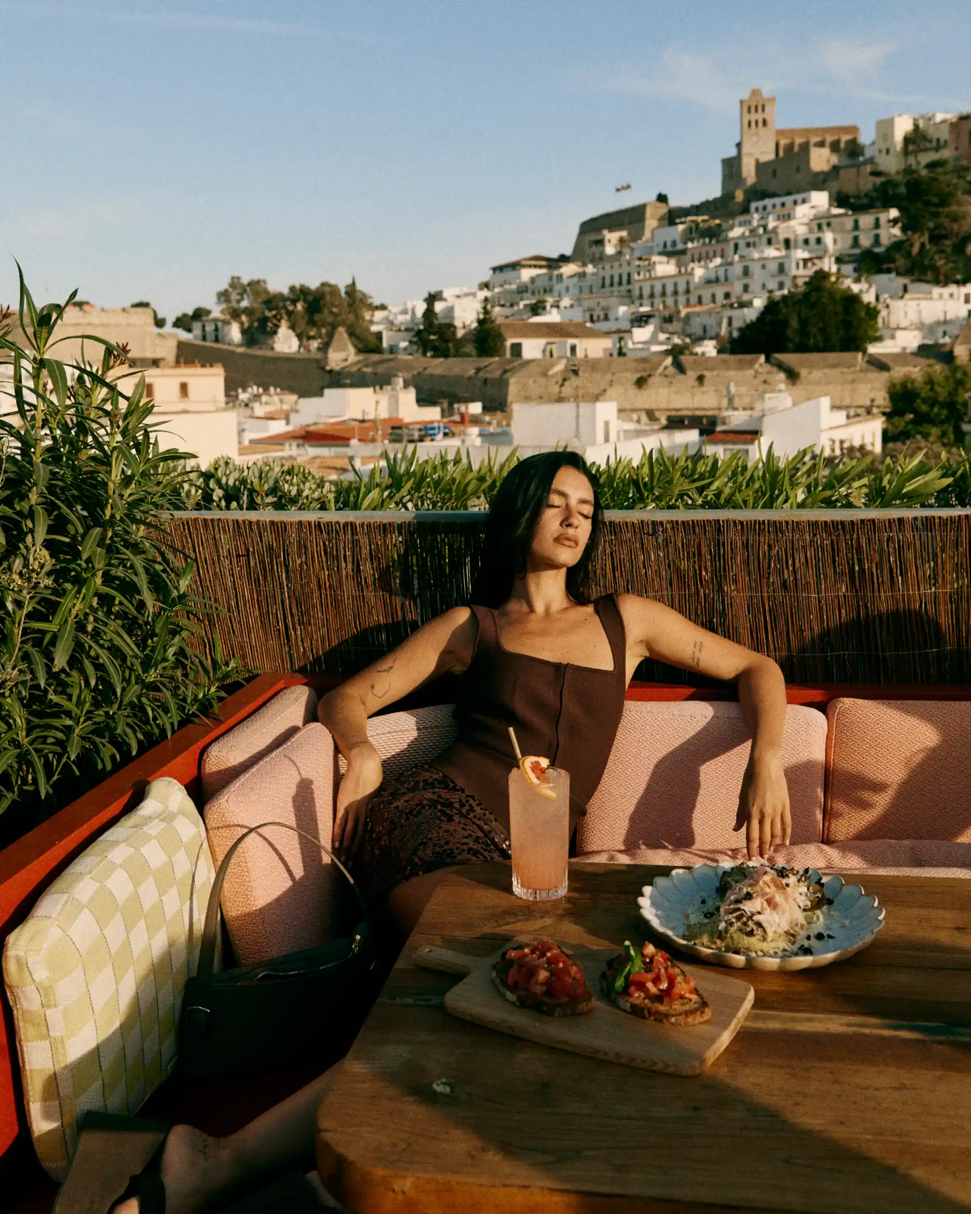 Woman in a dark dress relaxing on pink cushioned bench with food and drink on wooden table.