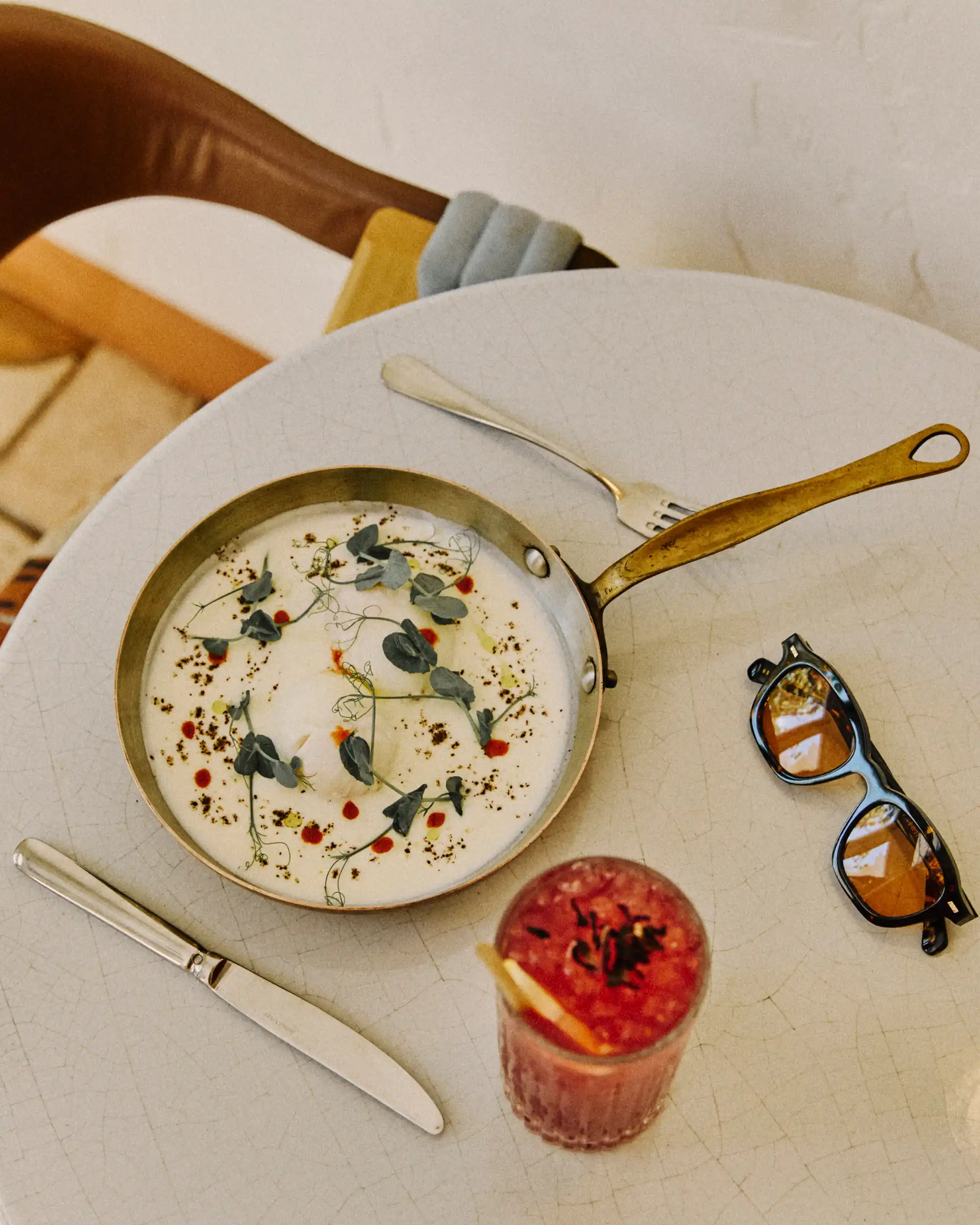 Table with small pan of food garnished with herbs, knife, fork, sunglasses, and a pink drink with a lemon slice.