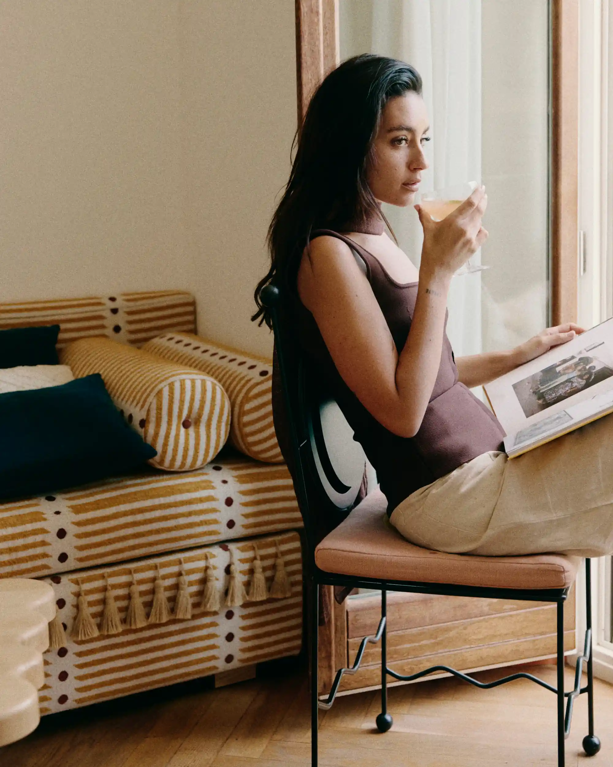 Woman sitting by a window on a chair drinking from a glass and reading a book.