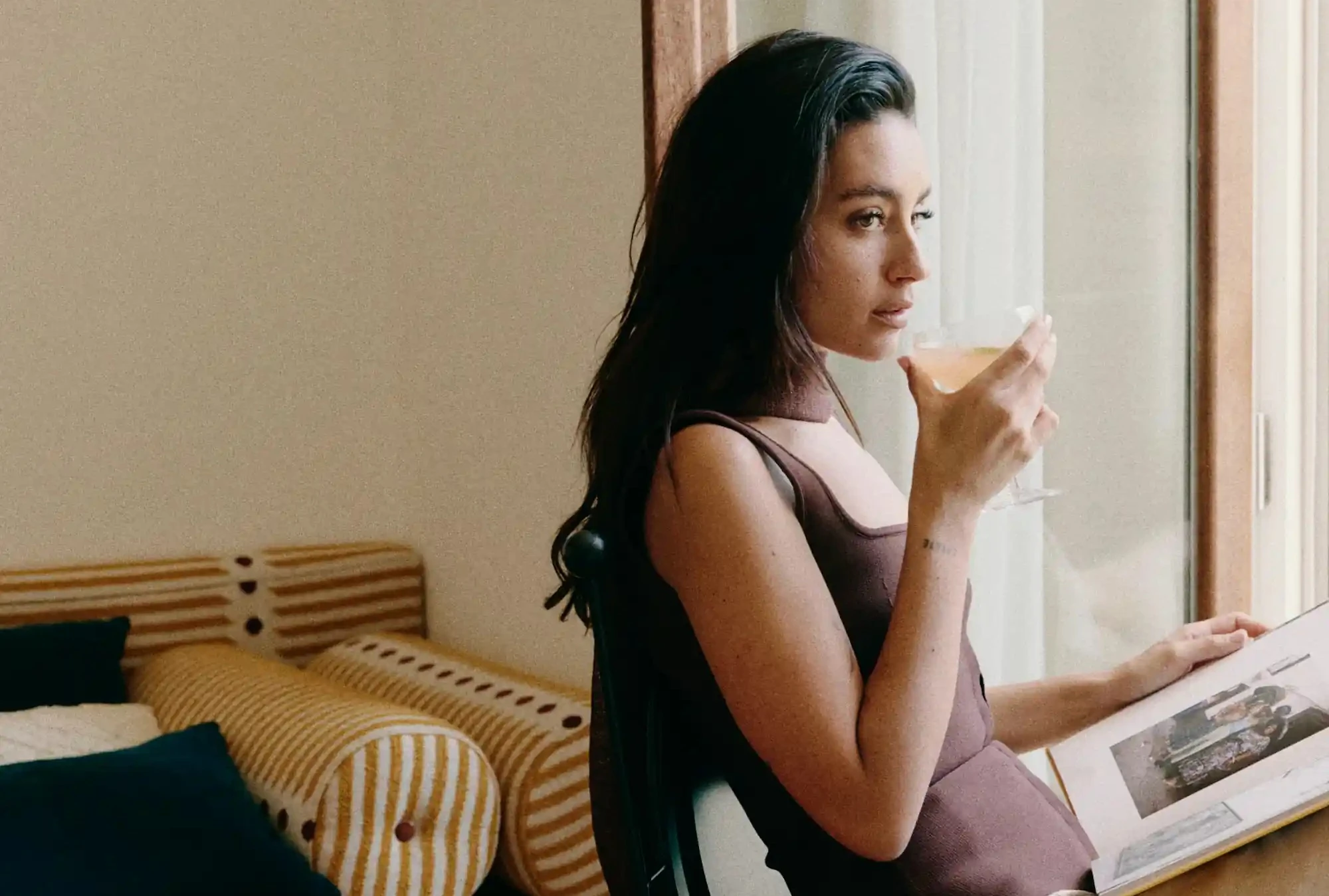 Woman in brown dress sitting by window, drinking from glass and looking away while holding a photo album.