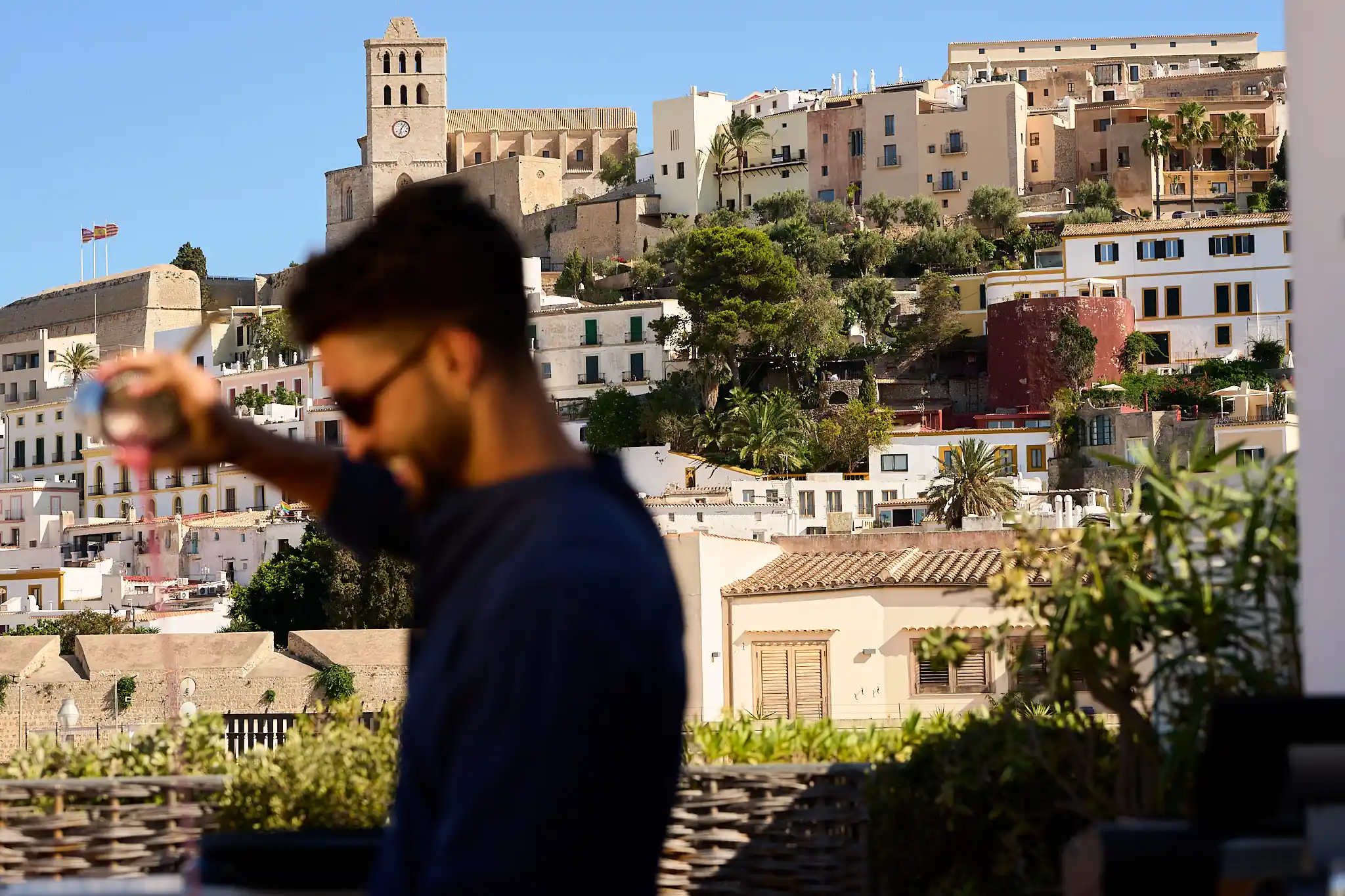 Blurred man in sunglasses with historic stone buildings and clock tower in background under blue sky.