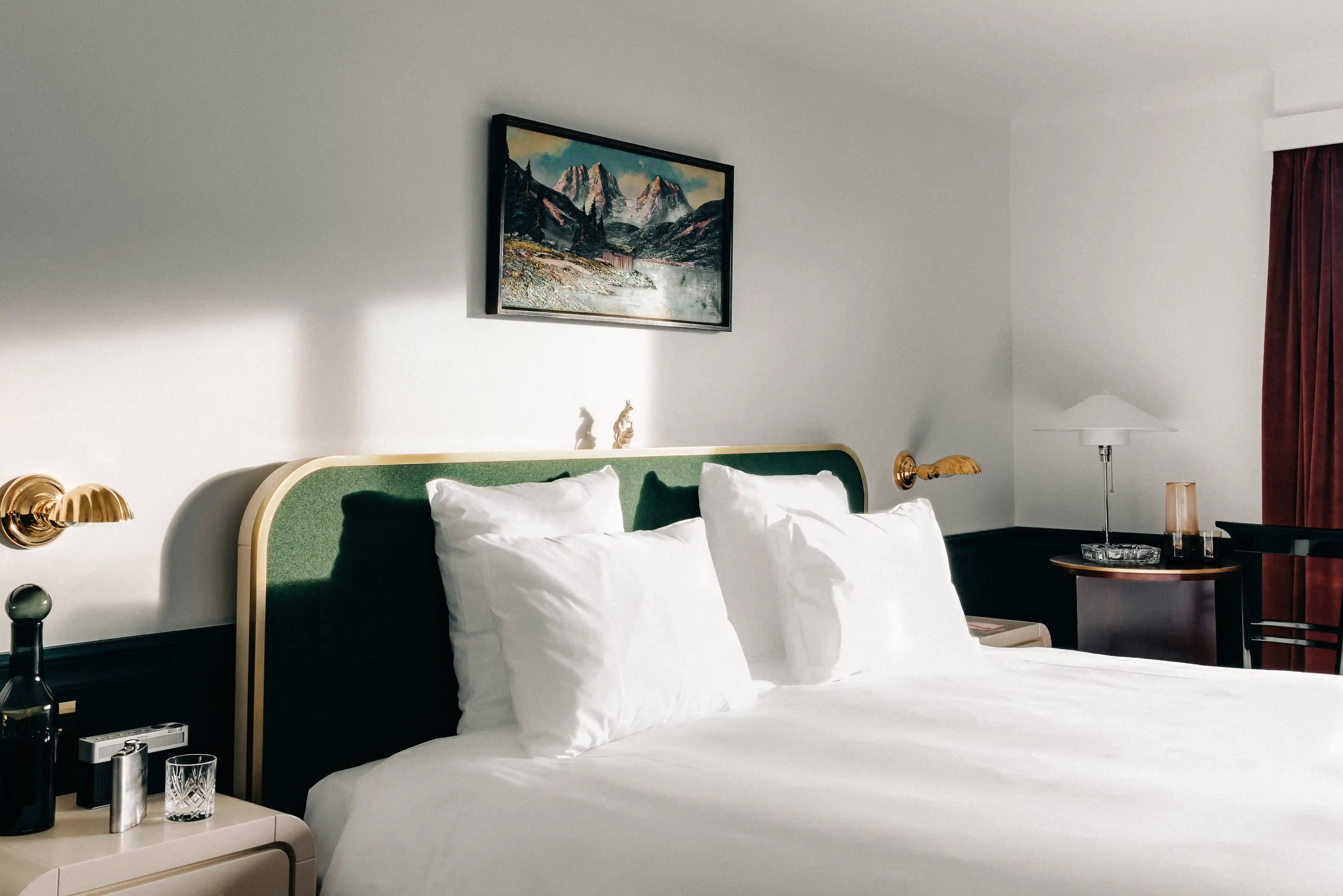 Sunlit bedroom with a bed with white pillows and green headboard beneath a mountain painting.