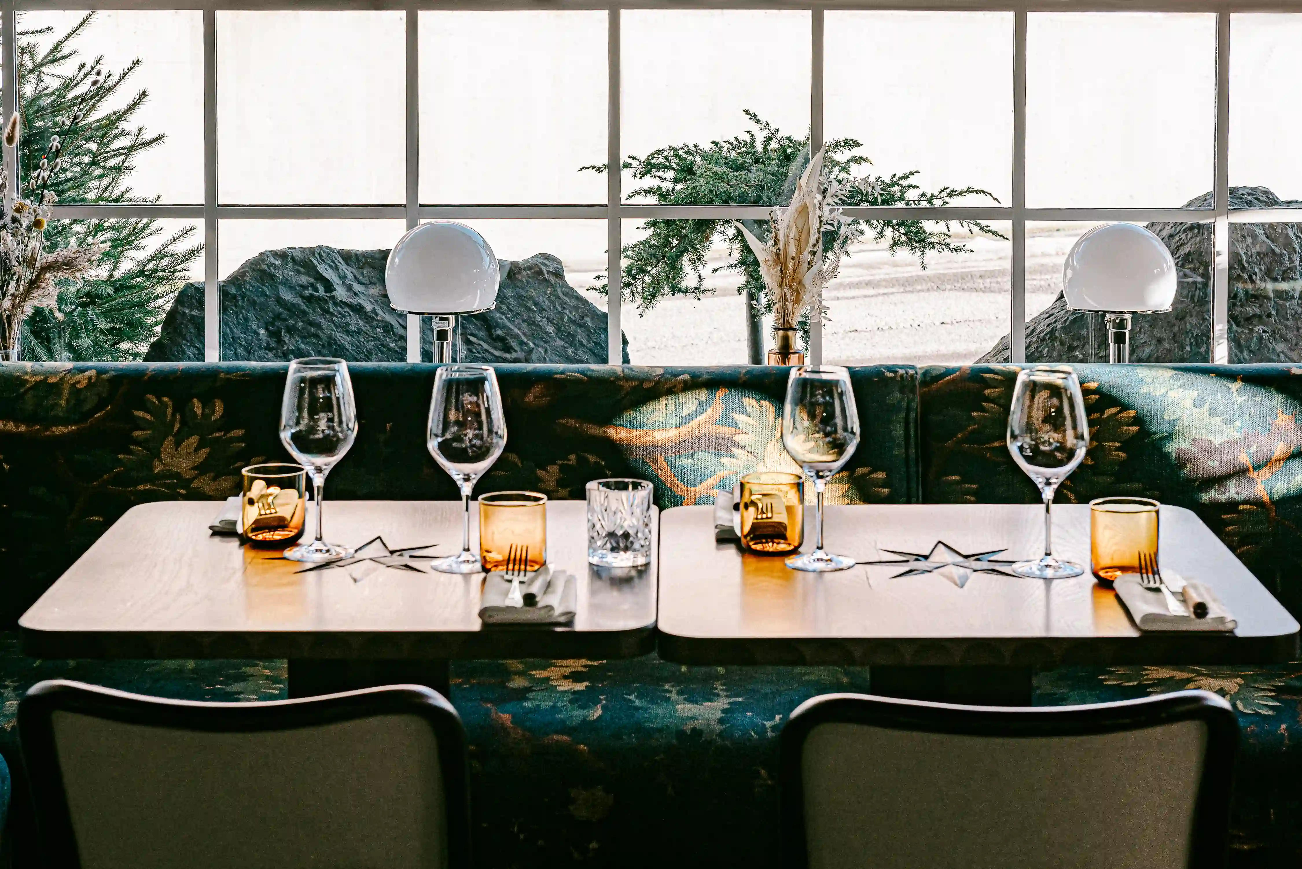 Two wooden tables set with wine glasses, amber glasses, and utensils in front of a window and patterned bench.