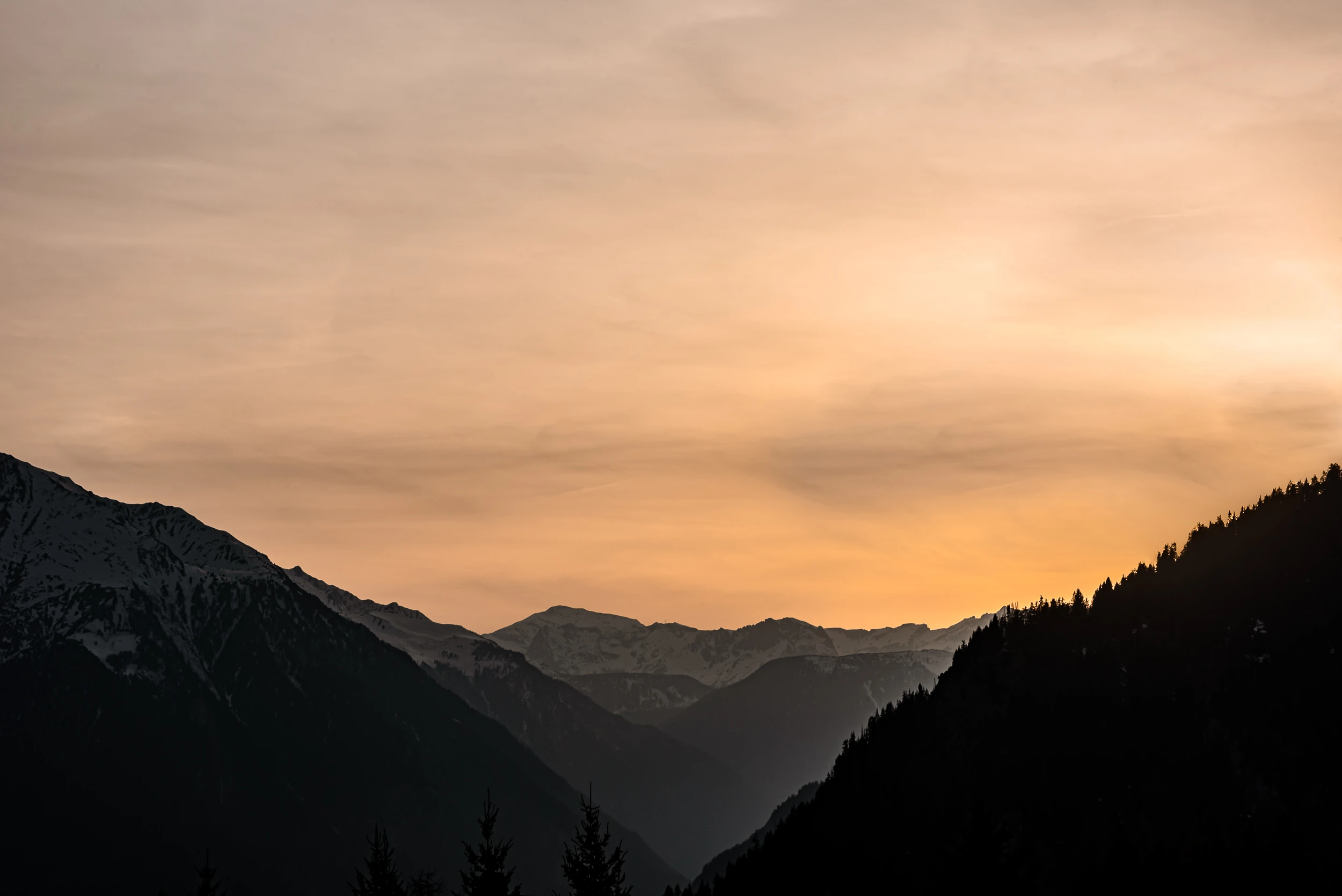 Silhouetted mountain ridges at sunset with soft orange and gray sky above snow-covered peaks.