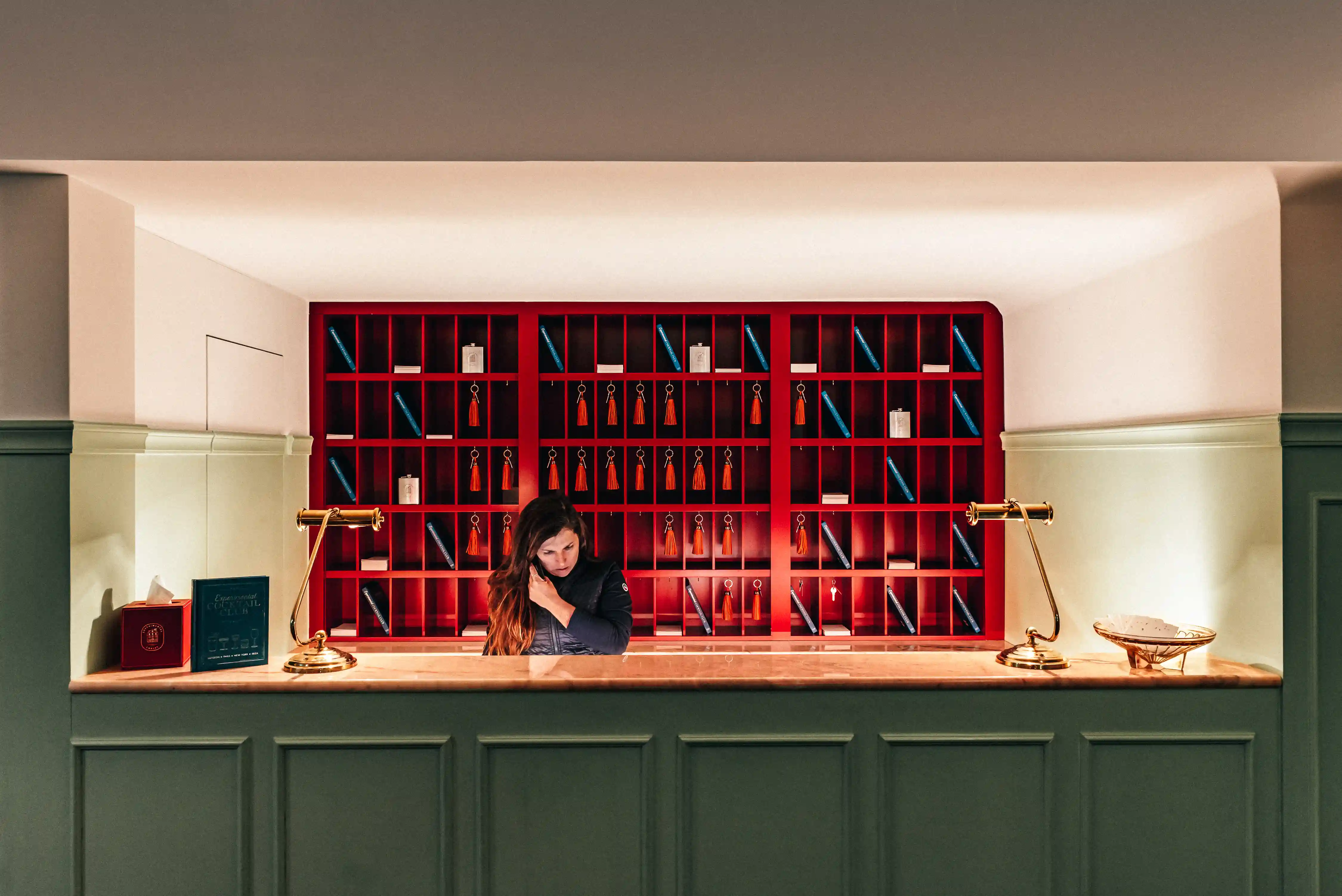 Reception desk with green paneling, two gold lamps, and a woman behind a red cubby wall with keys.