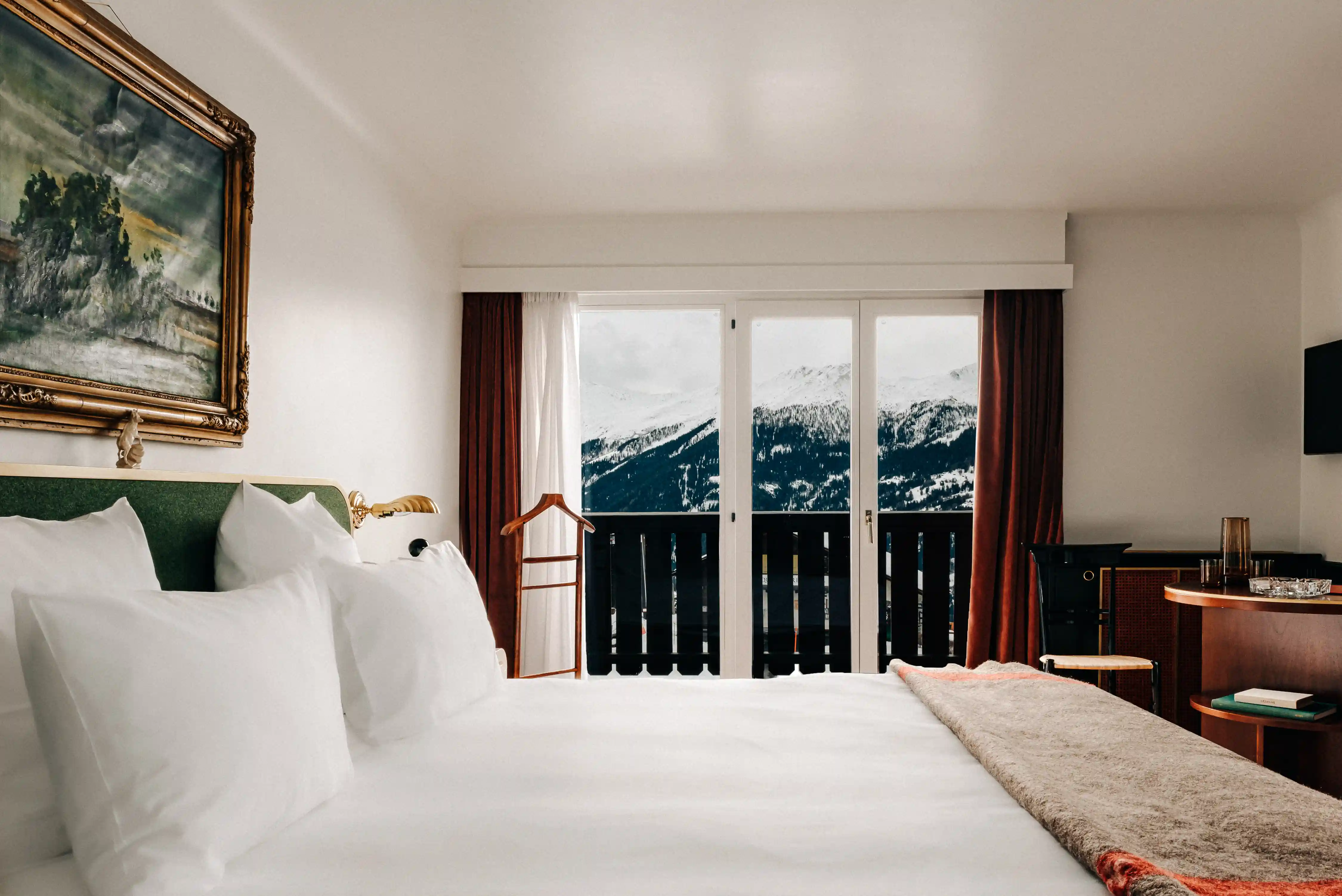 Cozy hotel room with white bedding, bedside lamps, painting, and snowy mountain view through glass doors.