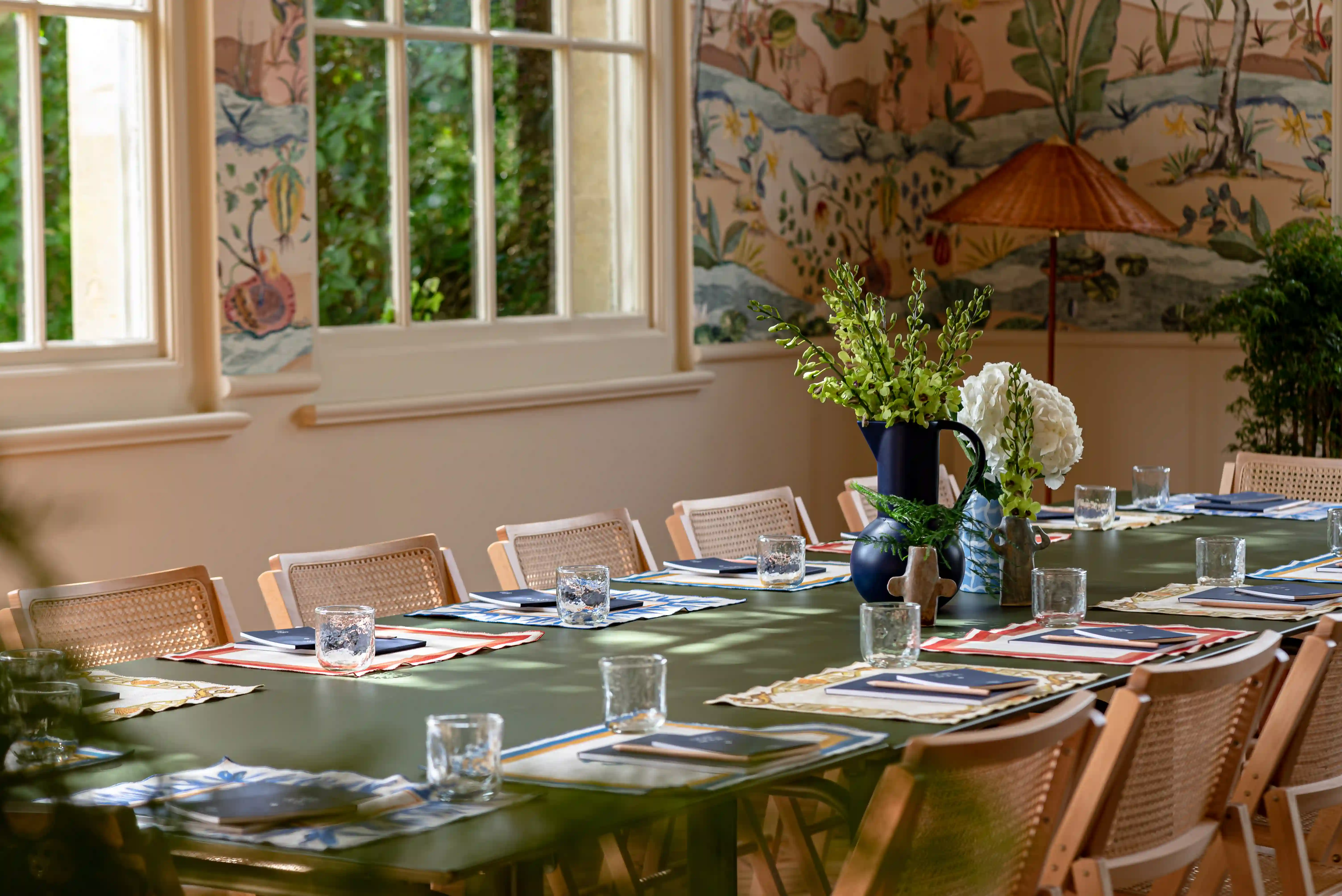 Long dining table set with colorful placemats, clear glasses, notebooks, and flower arrangements in vases.