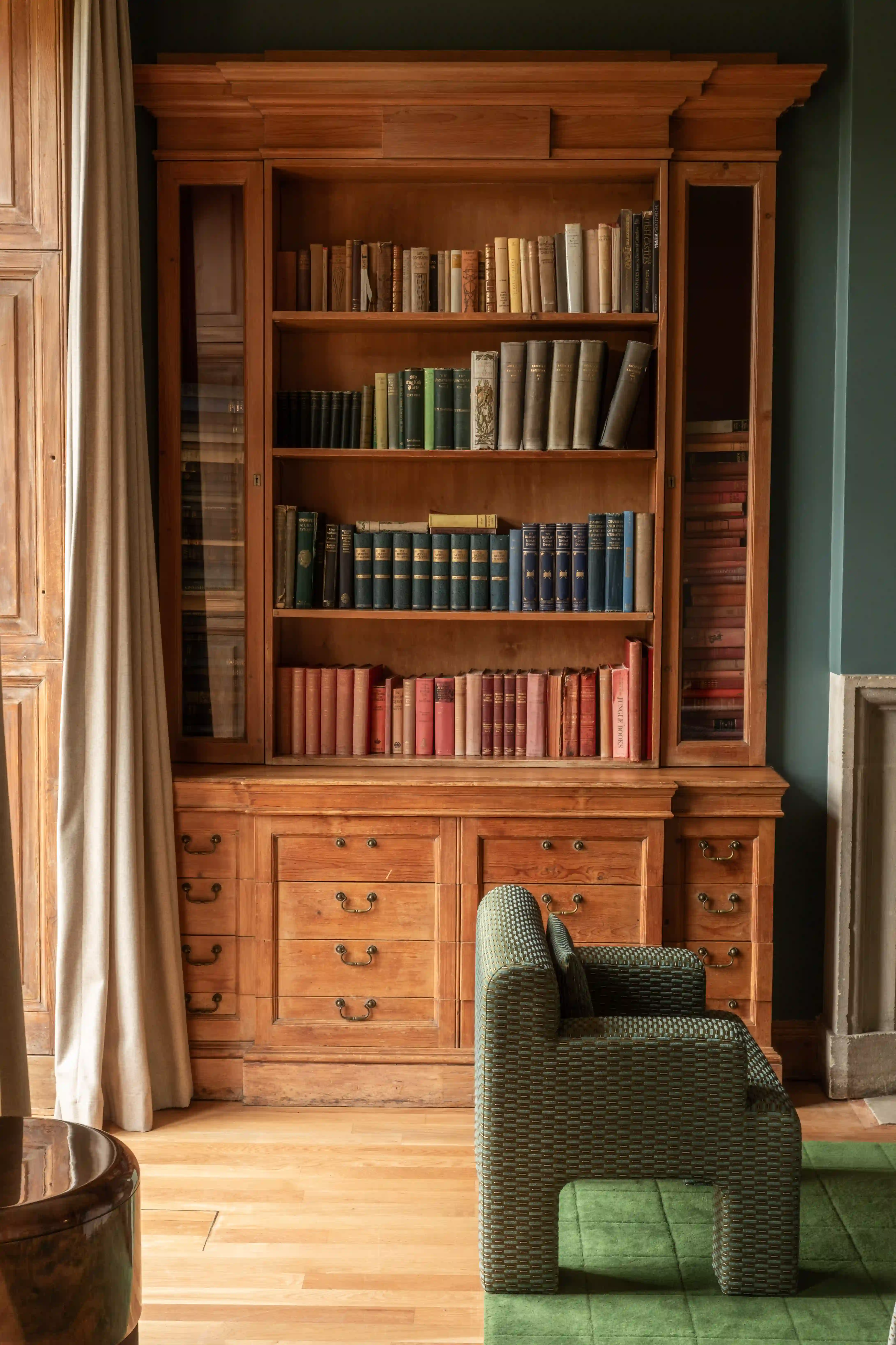 Wooden bookshelf filled with books above wooden drawers next to a green upholstered chair and curtains.