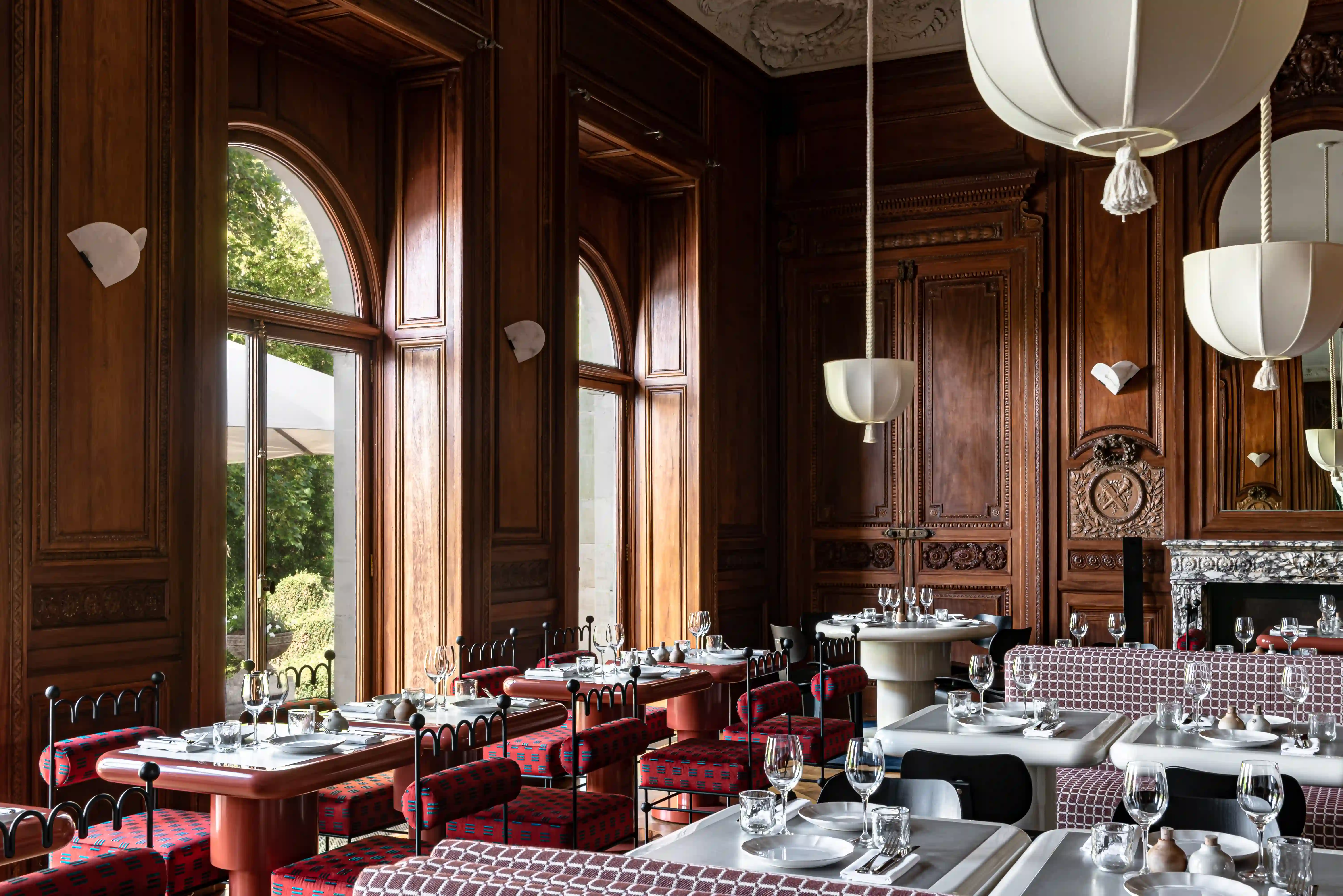 Elegant restaurant interior with wooden paneling, arched windows, red patterned chairs, and set dining tables.