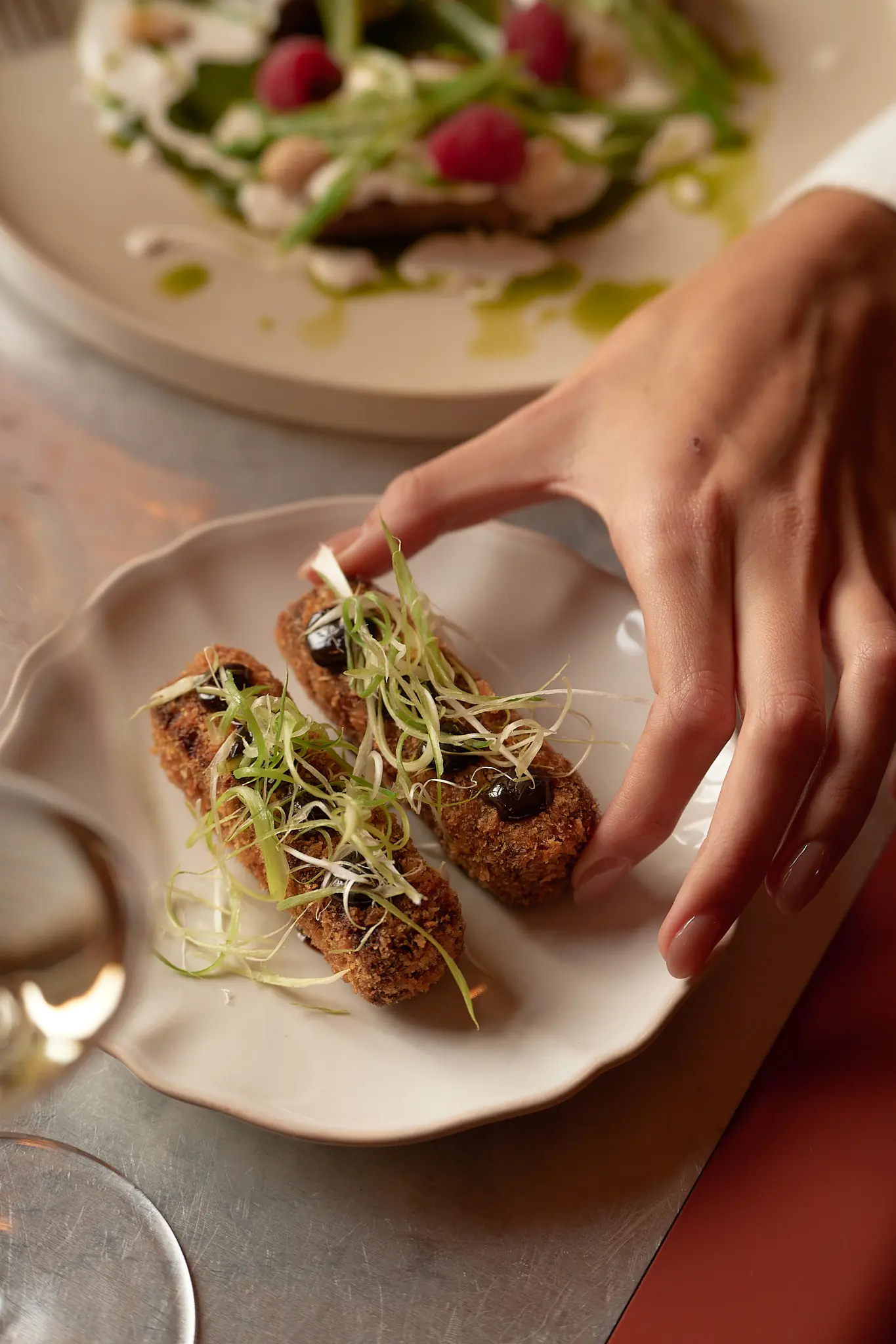 Hand picking a fried croquette topped with herbs and sauce from a white plate on a metal table.