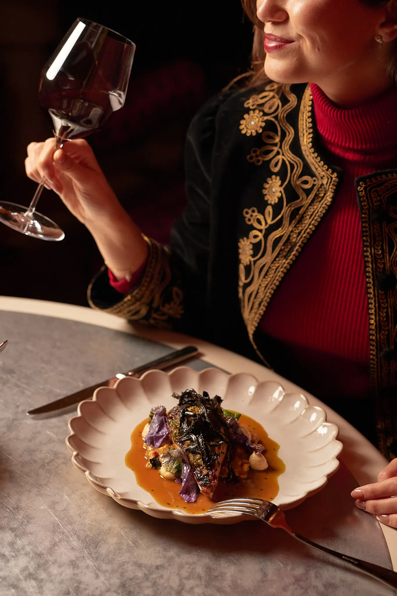 Woman in embroidered jacket holding a wine glass, with a gourmet meal on a scalloped plate.