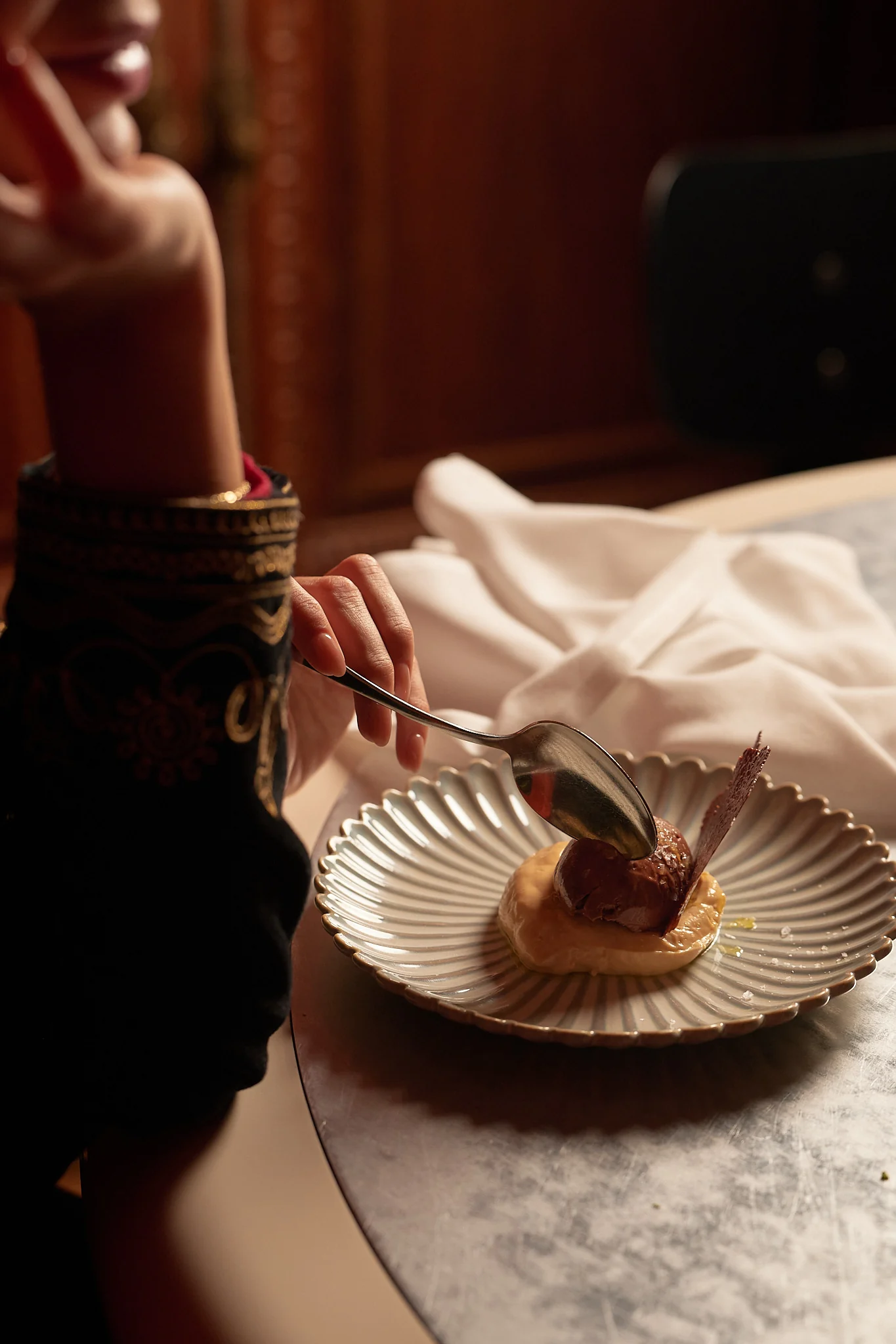 Person holding a spoon, about to eat a chocolate dessert on a decorative plate at a table.