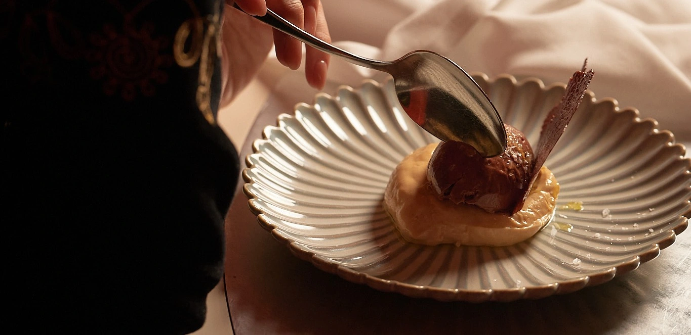 Hand holding a spoon scooping chocolate dessert on cream with decorative crisp on a fluted plate.