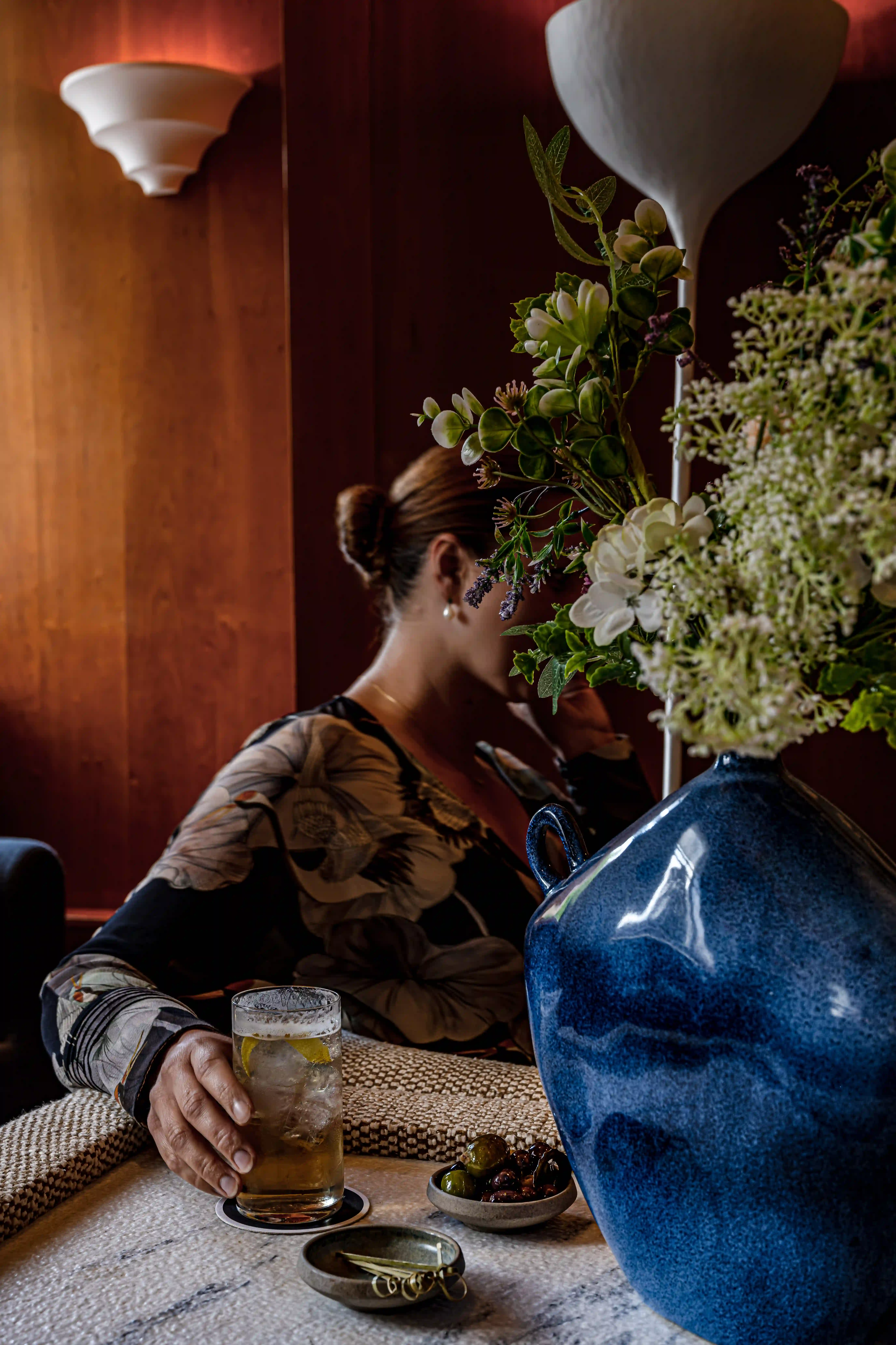 Woman sitting at a table with a drink and snacks, partially obscured by a large blue vase with flowers.