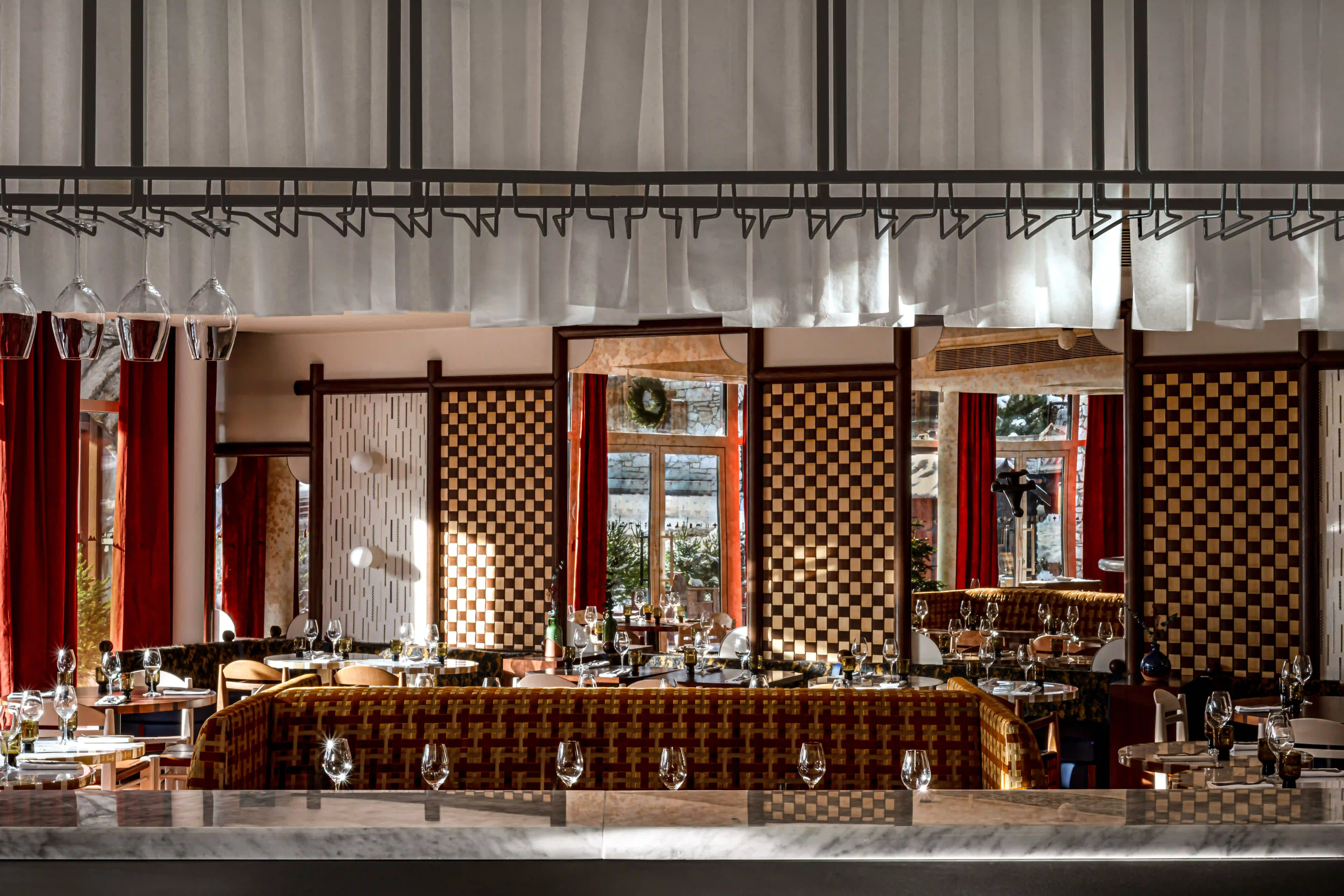 Upscale restaurant interior with patterned booths, checkered panels, wine glasses, and hanging glasses above a marble bar.