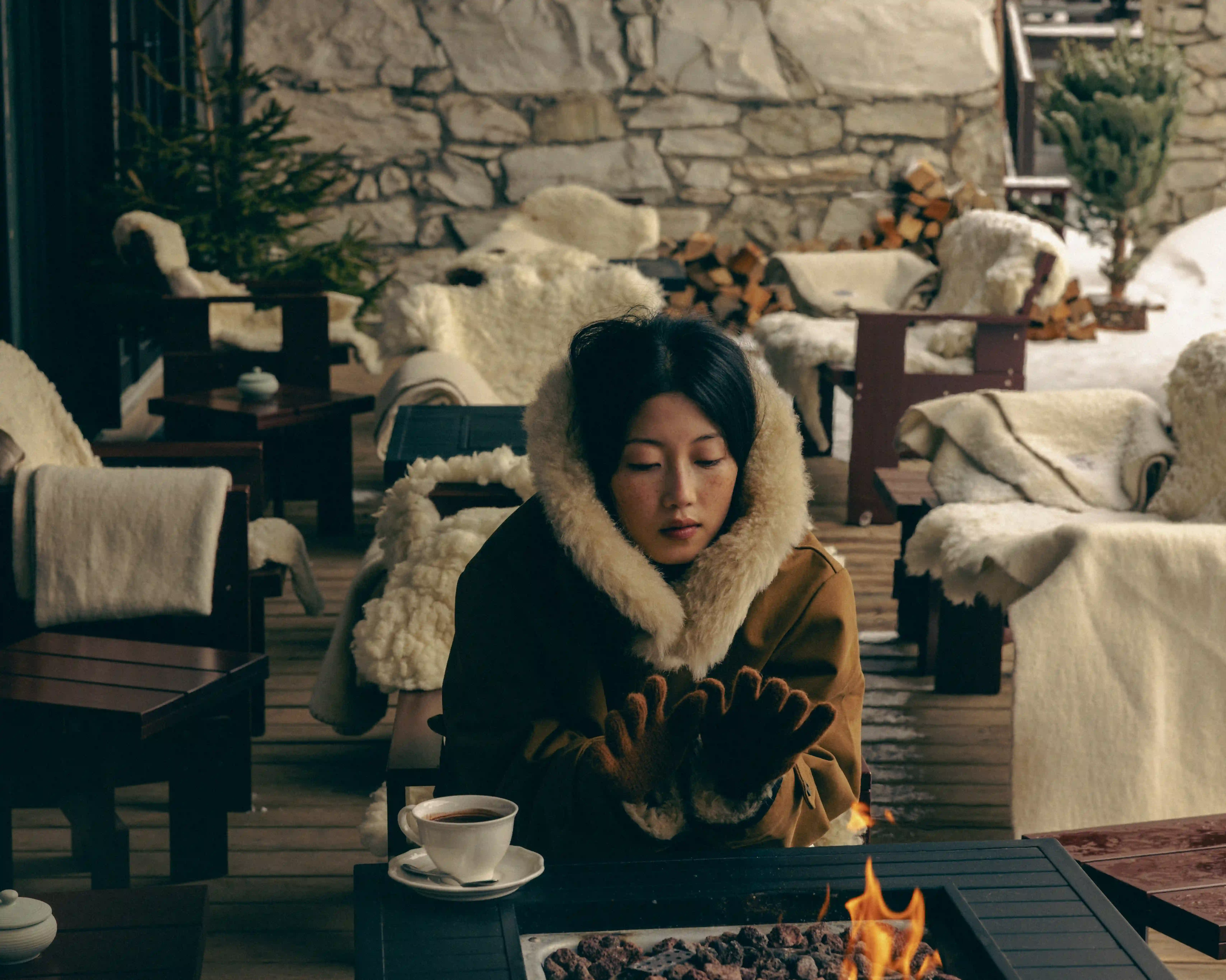 Woman in warm coat and gloves warming hands by fire pit on a cushioned outdoor patio in winter.