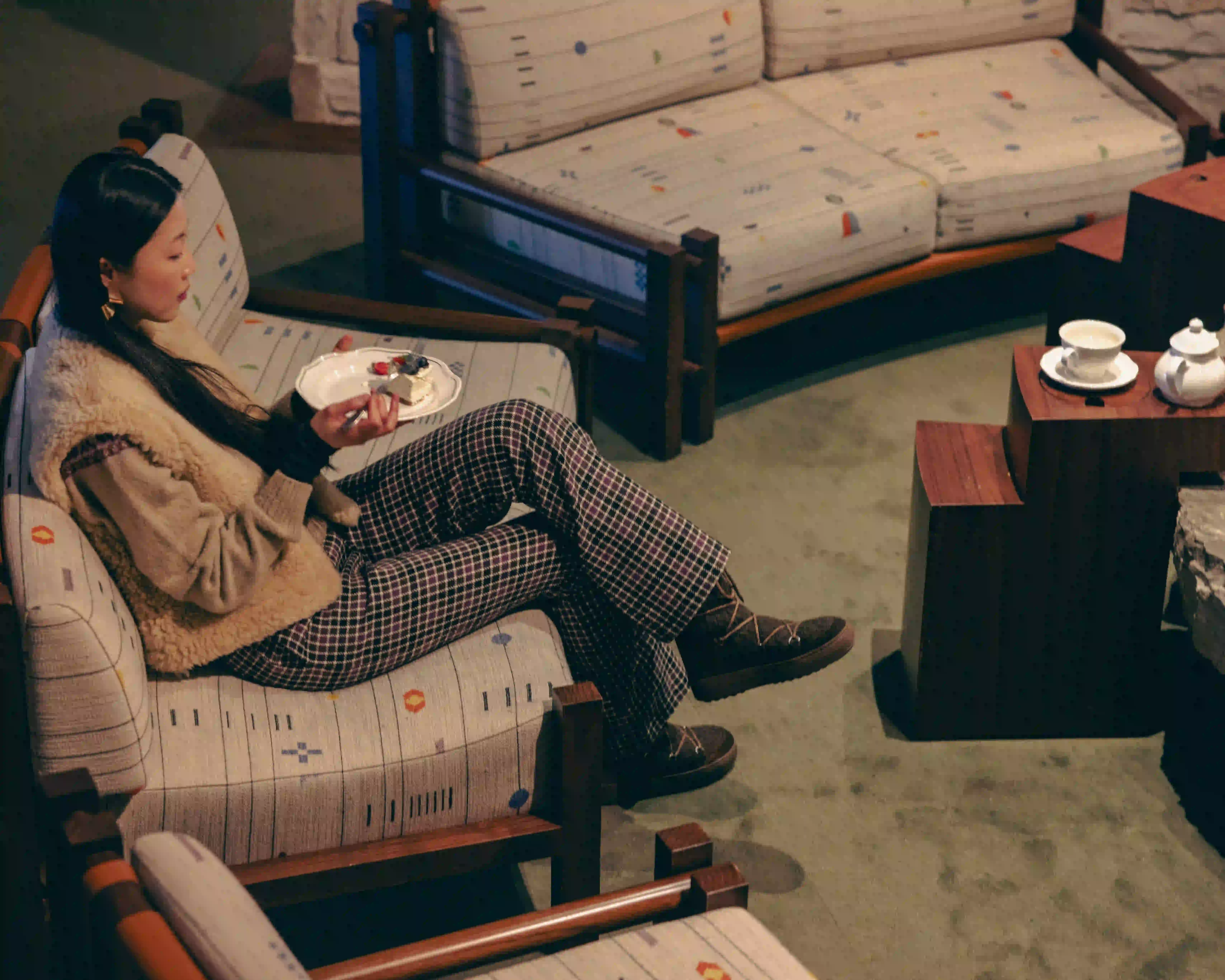 A woman in a fur vest sits on a patterned sofa eating cake in a cozy room with wooden furniture.