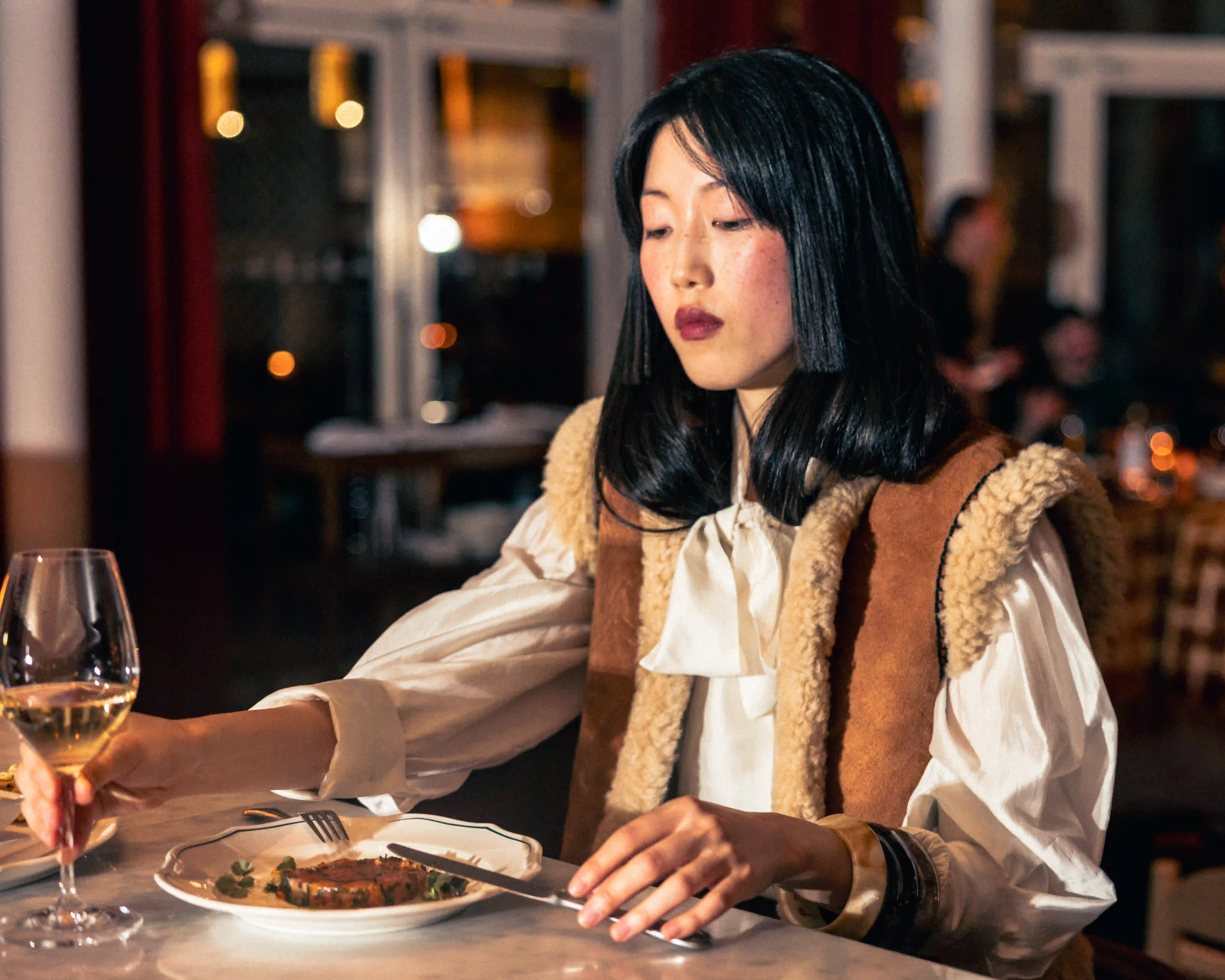 Woman with dark hair holding a glass of white wine and a knife at a dining table with food.