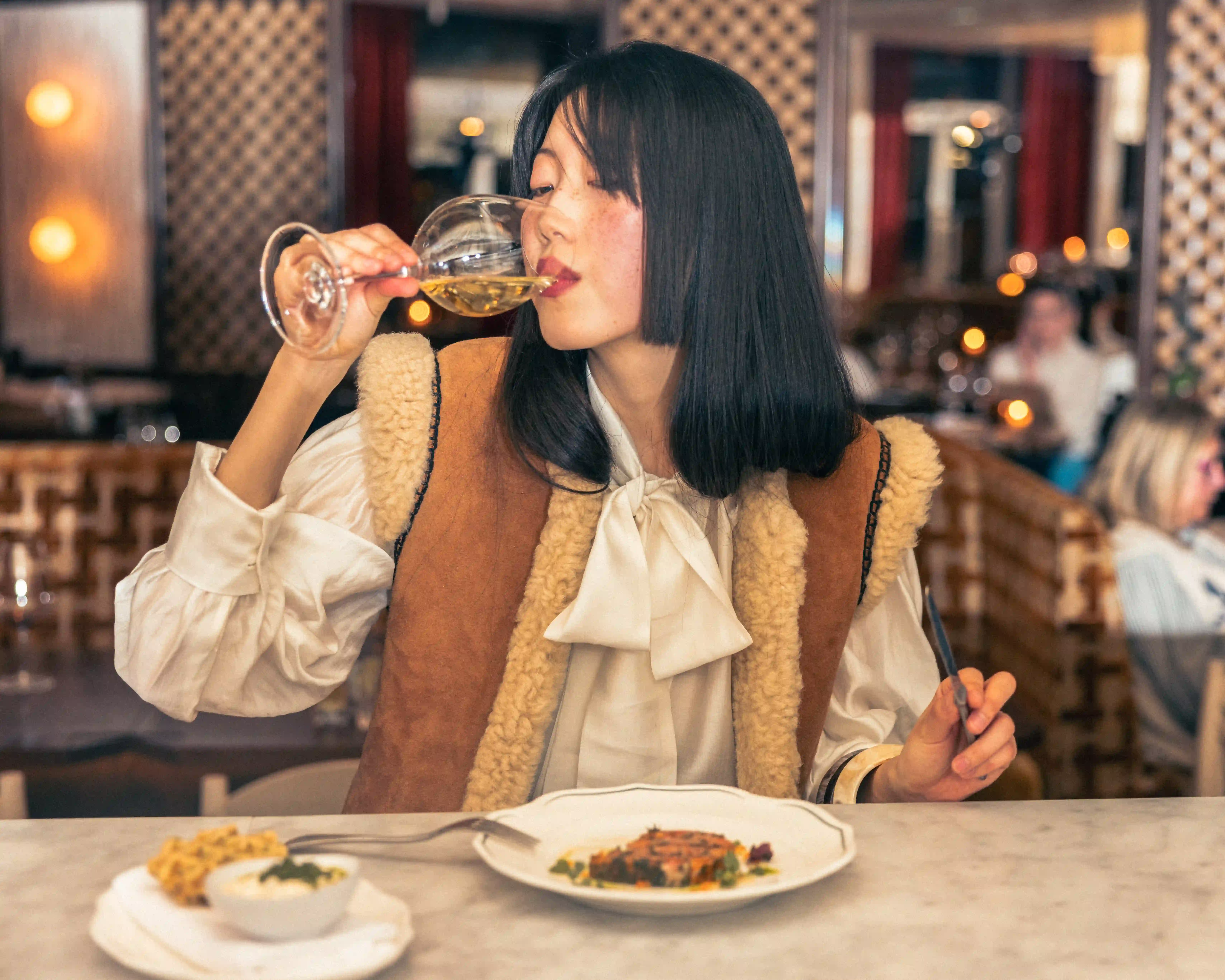 Woman with black hair drinking white wine at a restaurant table with food in front of her.