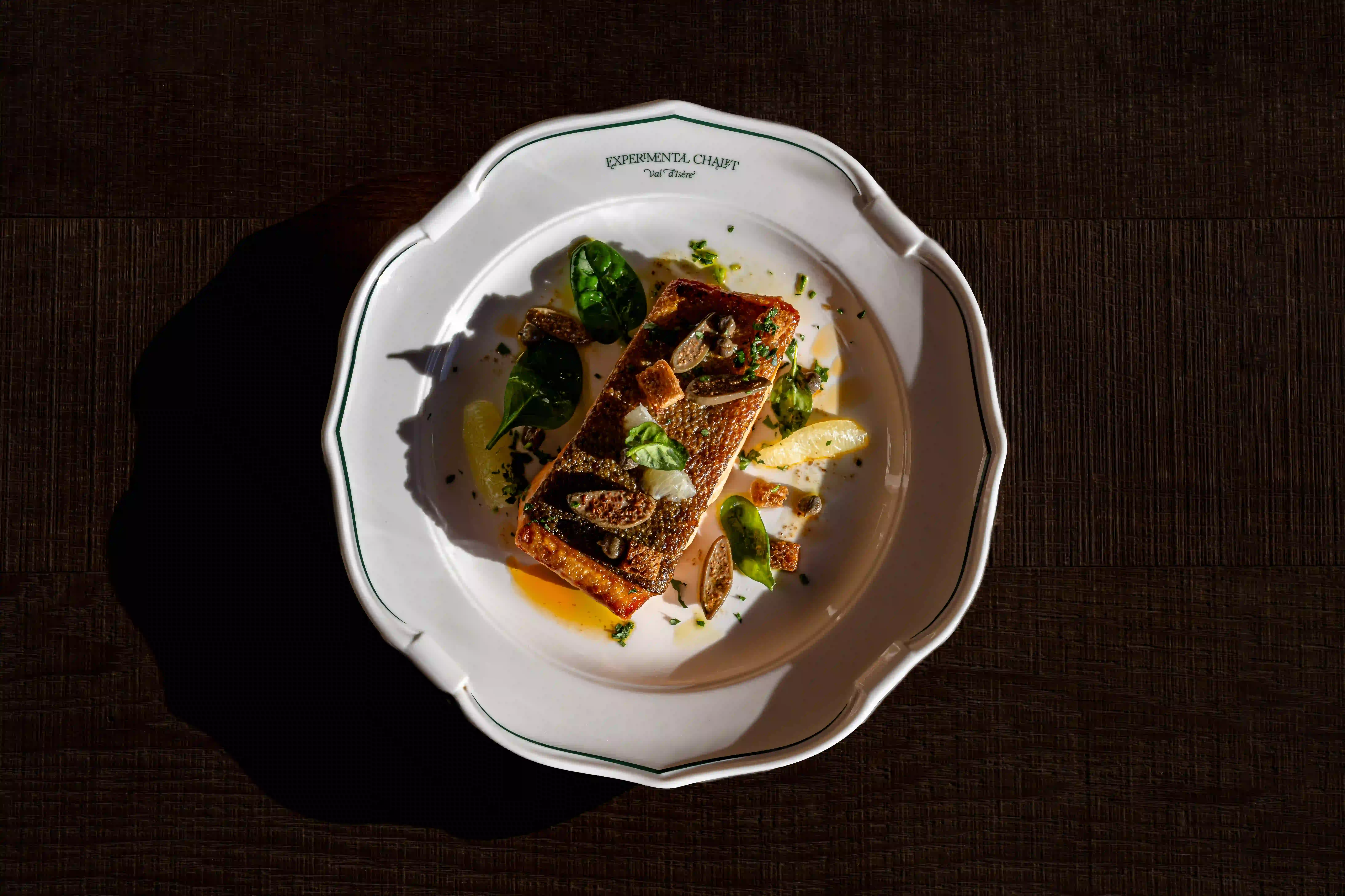 Seared fish fillet garnished with greens and citrus segments on a white decorative plate.