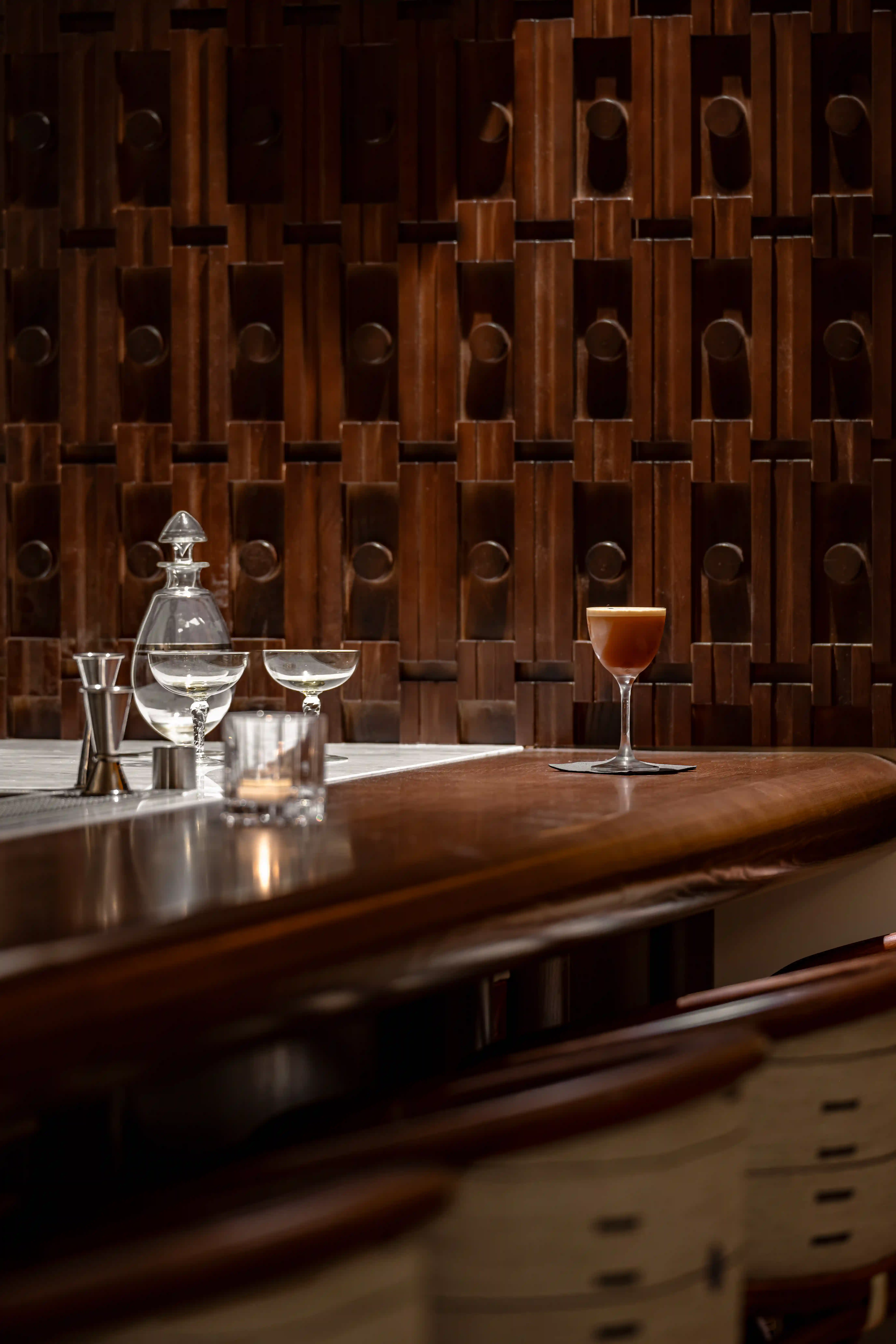Craft cocktail on wooden bar counter with glassware and textured wooden wall in dim light.