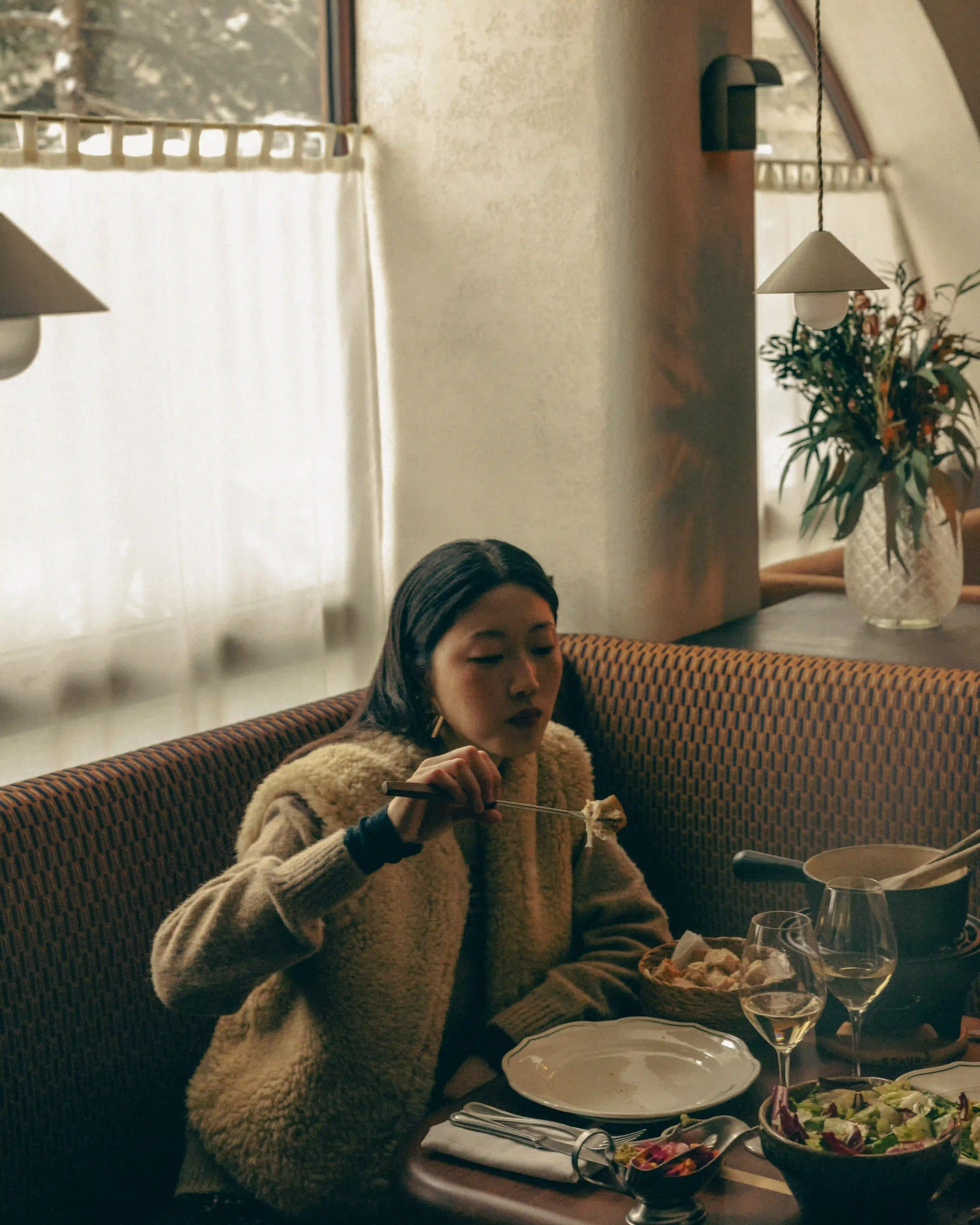 Woman in a cozy sweater eating fondue at a table with wine glasses and a salad bowl indoors.