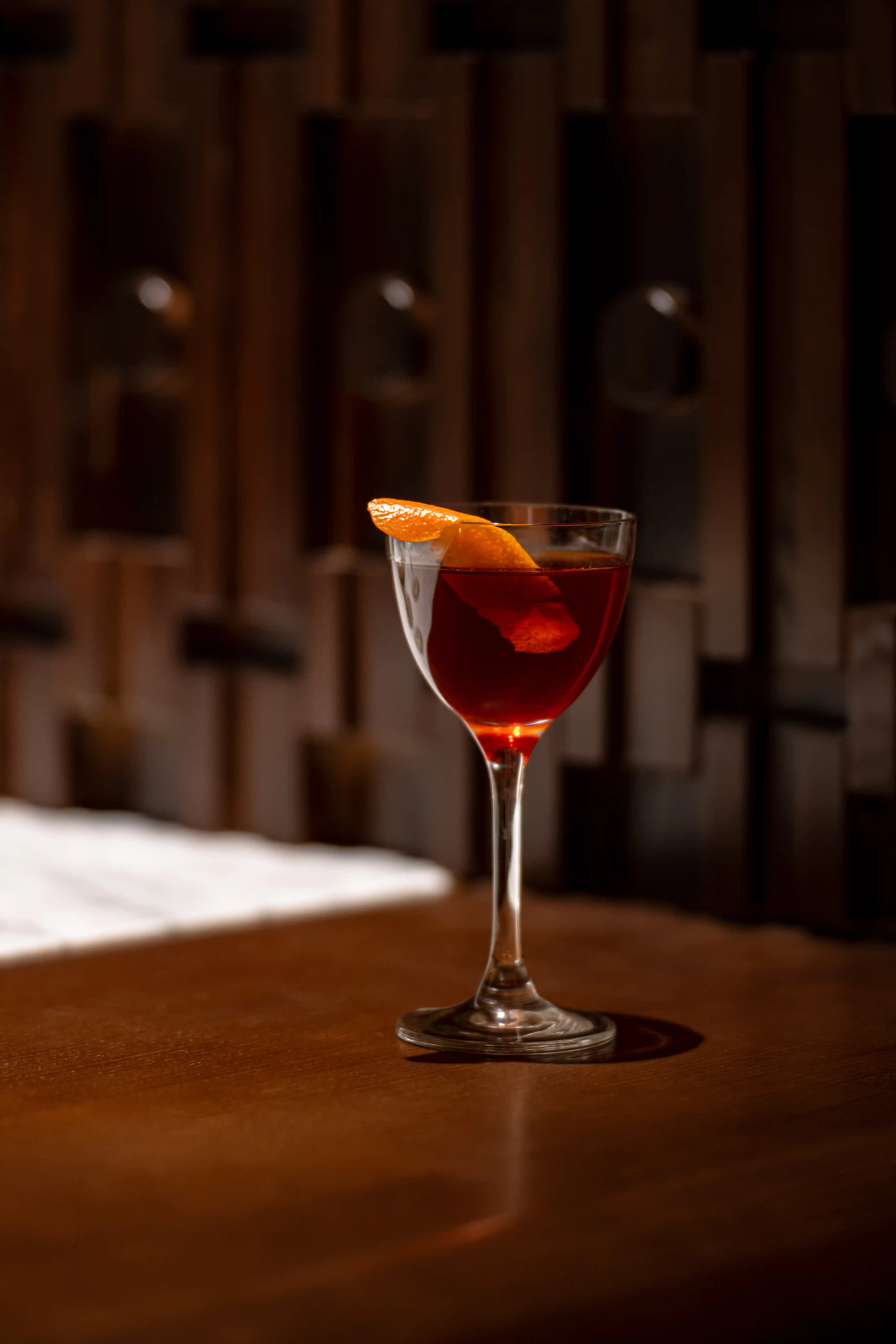 Cocktail glass with a dark red drink garnished with an orange peel on a wooden table.