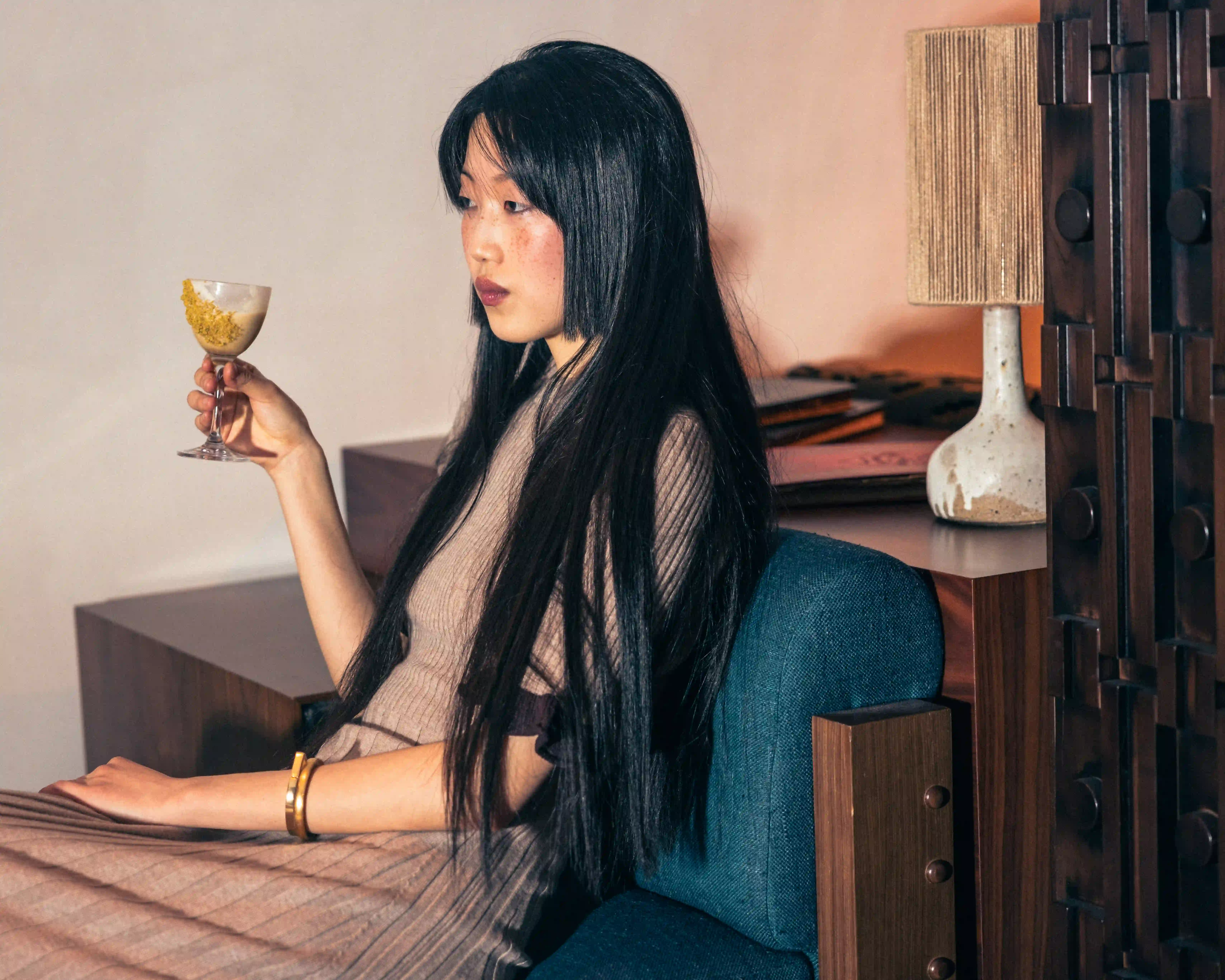 Woman with long black hair sitting on a blue chair holding a glass with yellow garnish.
