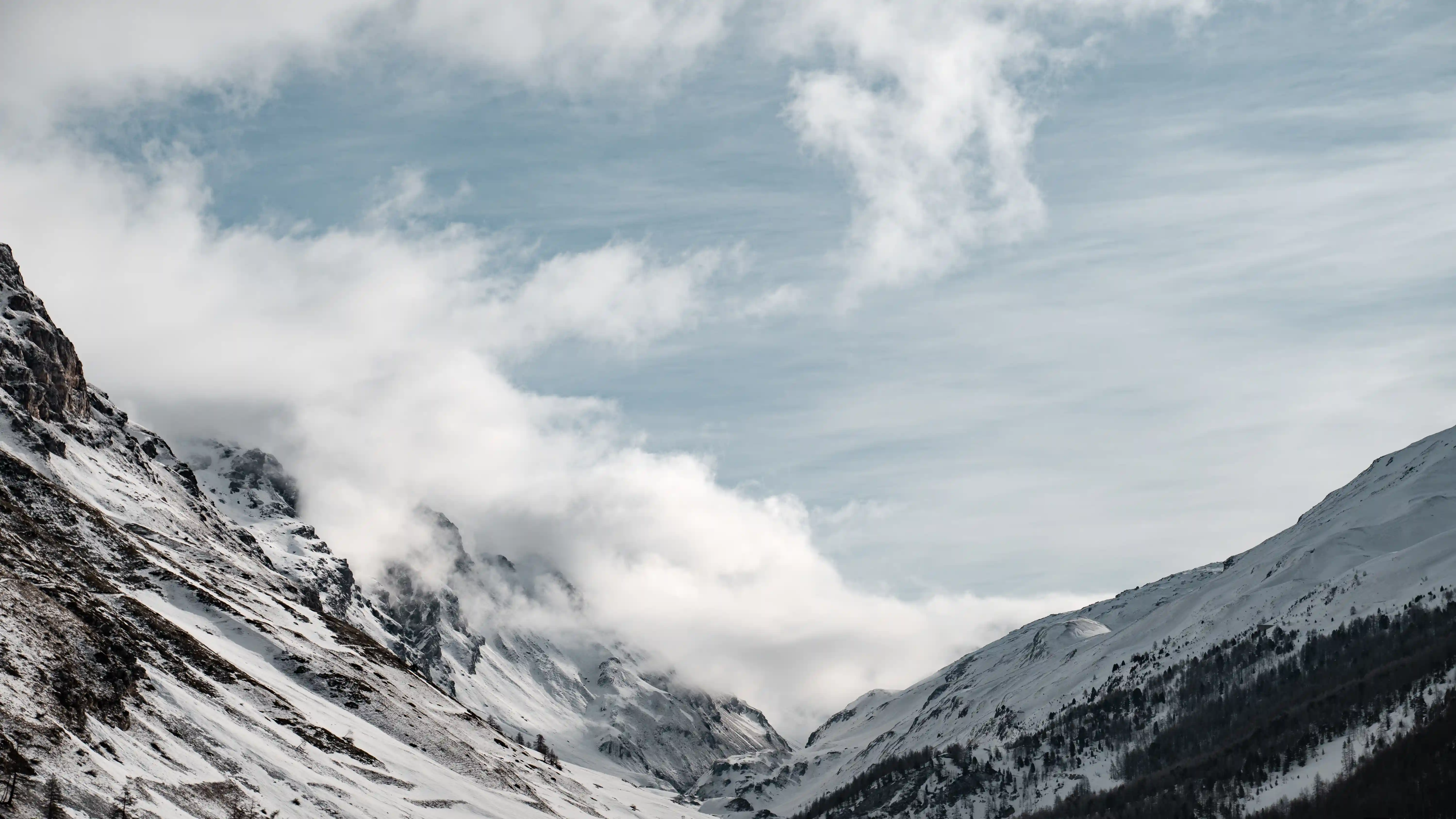 Snow-covered mountain valley with clouds drifting between the peaks under a blue sky.